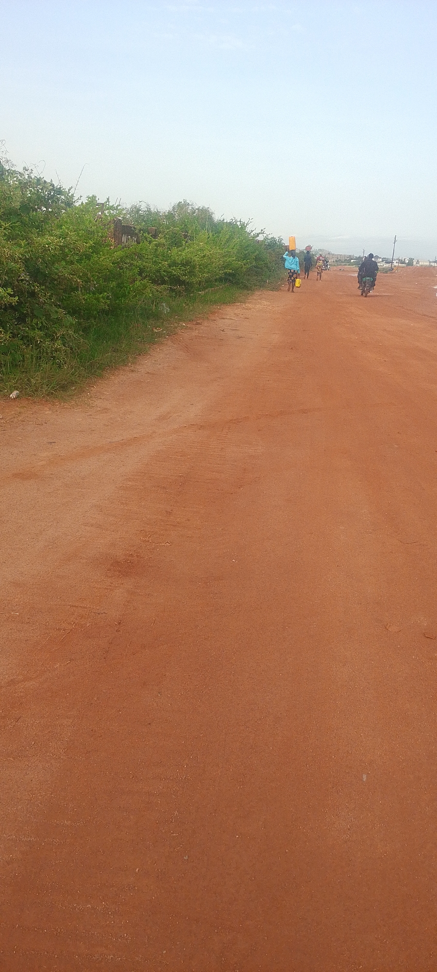 The image depicts a dusty, reddish-brown dirt road in Kwang, Nigeria. Green shrubbery lines the left side of the road. Several people are visible walking along the road, with one person carrying a yellow bucket on their head and another riding a motorcycle. The sky is a hazy, pale blue. The overall scene suggests a typical day in this rural Nigerian location.