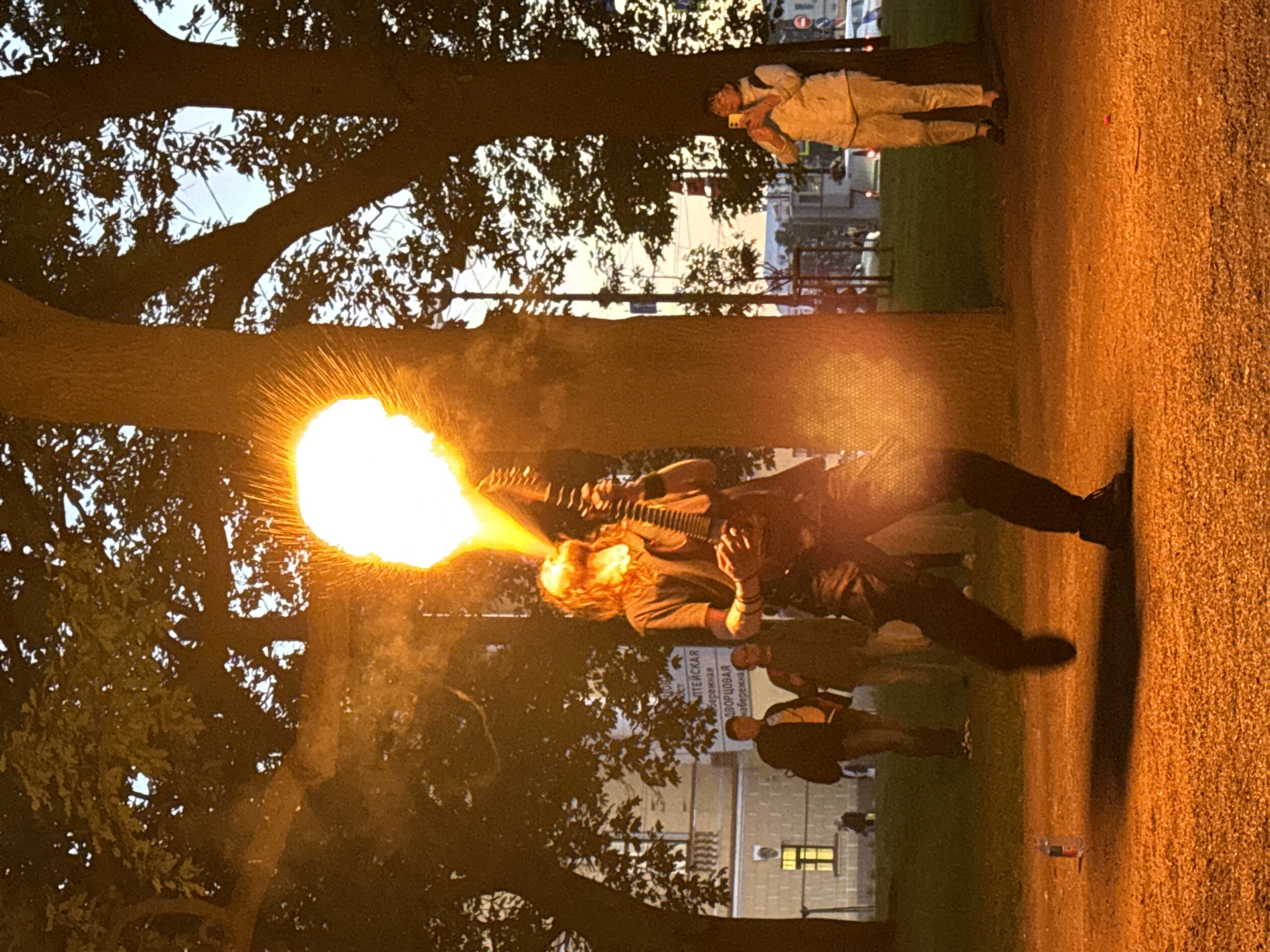 The image depicts a fire-breather performing with a flaming prop in front of a crowd in Saint Petersburg, Russia. The performer, dressed in dark clothing, is exhaling a large burst of fire while holding a musical instrument resembling a guitar. The performance is taking place outdoors, with large trees and a building in the background, suggesting a park or public square setting. Several spectators are visible, including a woman holding a phone to record the event. The ground is covered in gravel and illuminated by the fire's glow. The overall scene suggests an evening event with a lively and dramatic atmosphere.