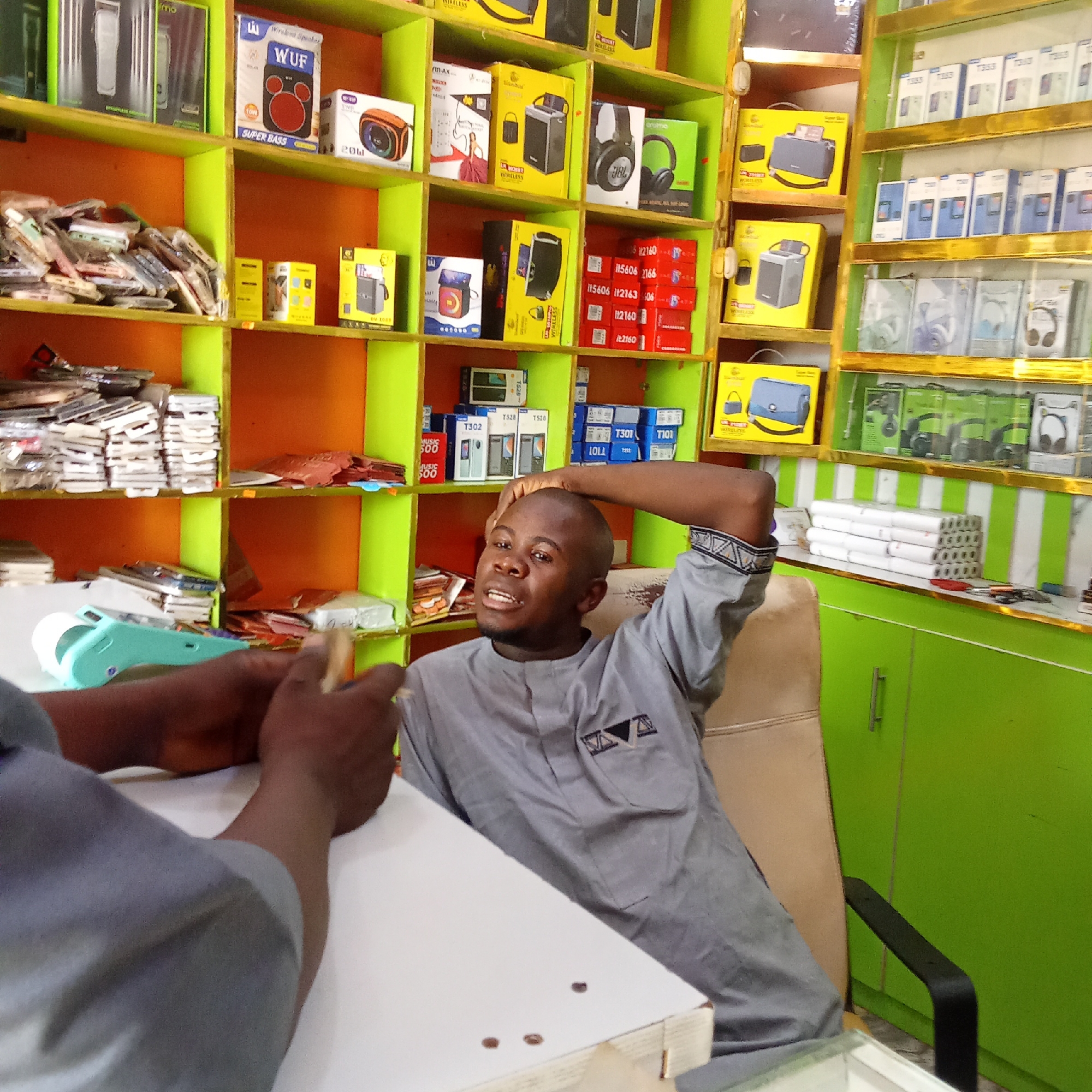 A photograph captures a man sitting in what appears to be a retail shop, likely selling mobile phones and accessories. The shelves behind him are stocked with a variety of items, including phone cases, speakers, headphones, and phone packaging. The shelves are painted orange and green, adding a vibrant but cluttered feel to the scene.
The man is dressed in a grey shirt with geometric patterns on the sleeves. He is seated in a tan-colored chair and has one arm resting behind his head. In the foreground, another person is partially visible, their arm and hand suggesting an interaction, perhaps in the context of a transaction or conversation within the shop.
The shop's layout is dense, with products displayed on shelves and a white countertop in the front. The lighting appears to be a mix of natural and artificial sources, illuminating the interior. The overall impression is that of a bustling, small-scale retail environment.