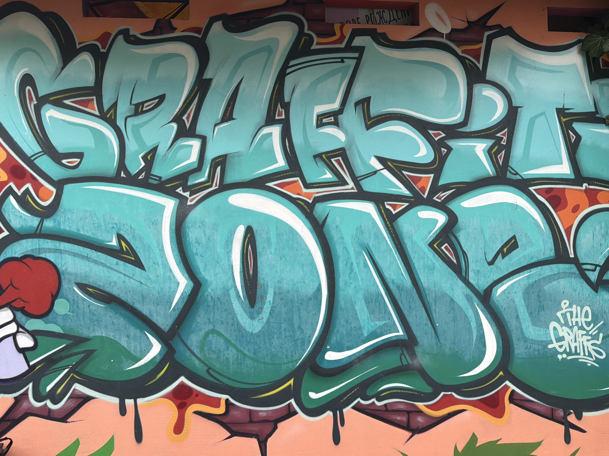 A color photograph shows graffiti on a wall. The graffiti is in a teal color, with dark green, white, black, orange, and red accents. The word "GRAFF SOA" is written across the center of the image. The artist's tag "THE GRAFFS" is in white in the lower right corner of the image. The wall appears to be made of peach and red brick and drips of dark green and black paint cascade down from the graffiti onto the brick. There are some dark shapes near the top of the image behind the graffiti. The location is Varna, Bulgaria.