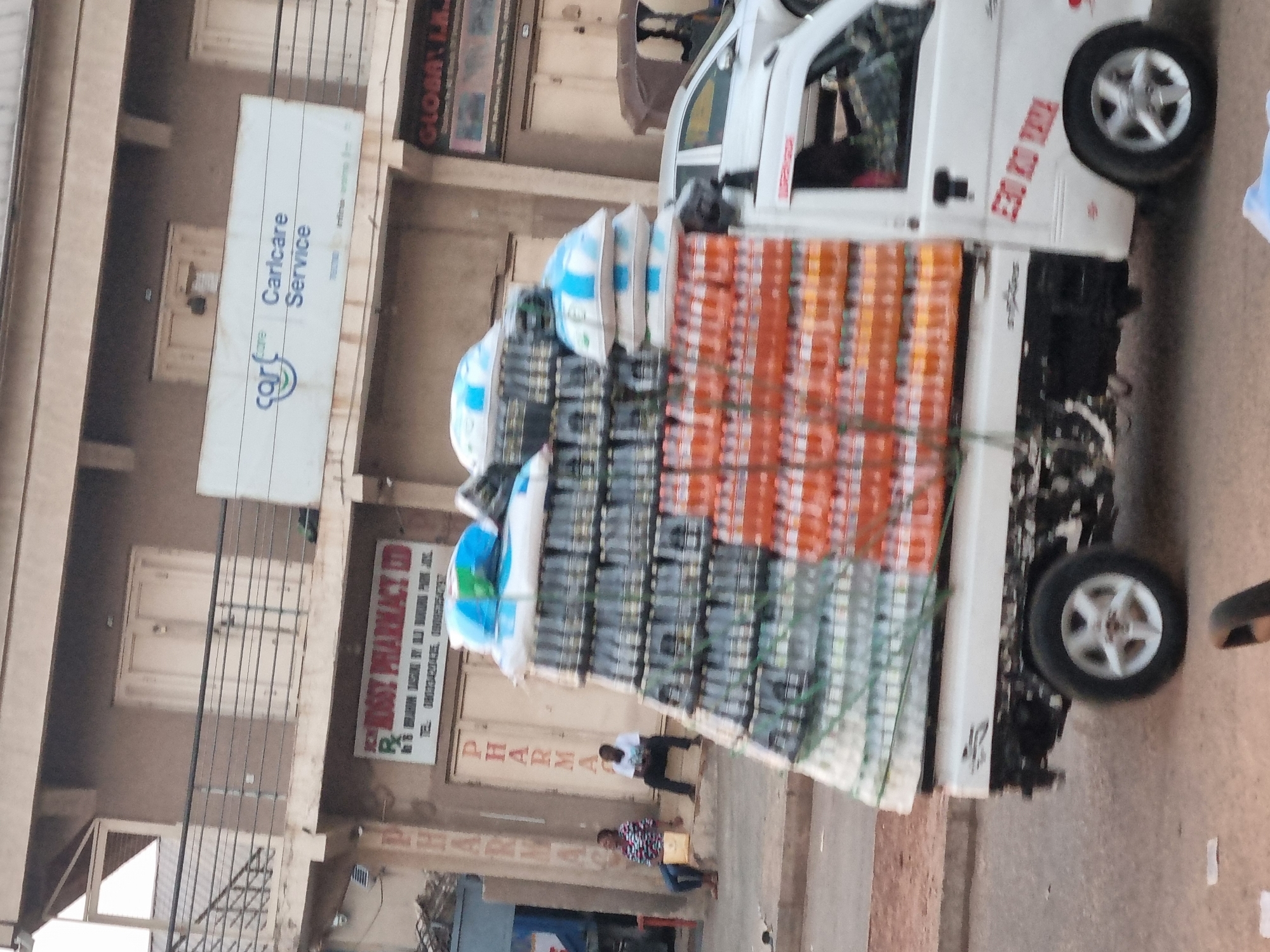 A daytime image depicts a white truck heavily laden with cargo, potentially goods for sale, parked on a street in Jos, Nigeria. The cargo appears to consist of stacked items wrapped in packaging, predominantly blue and orange hues. Two individuals are seated on the steps of a building nearby, appearing to observe the scene. The architecture suggests an urban environment. Visible signs include "Carlcare Service" and "Rose Pharmacy LTD," indicating the presence of commercial establishments in the vicinity. The truck displays the text "E20 KDA TRUK".