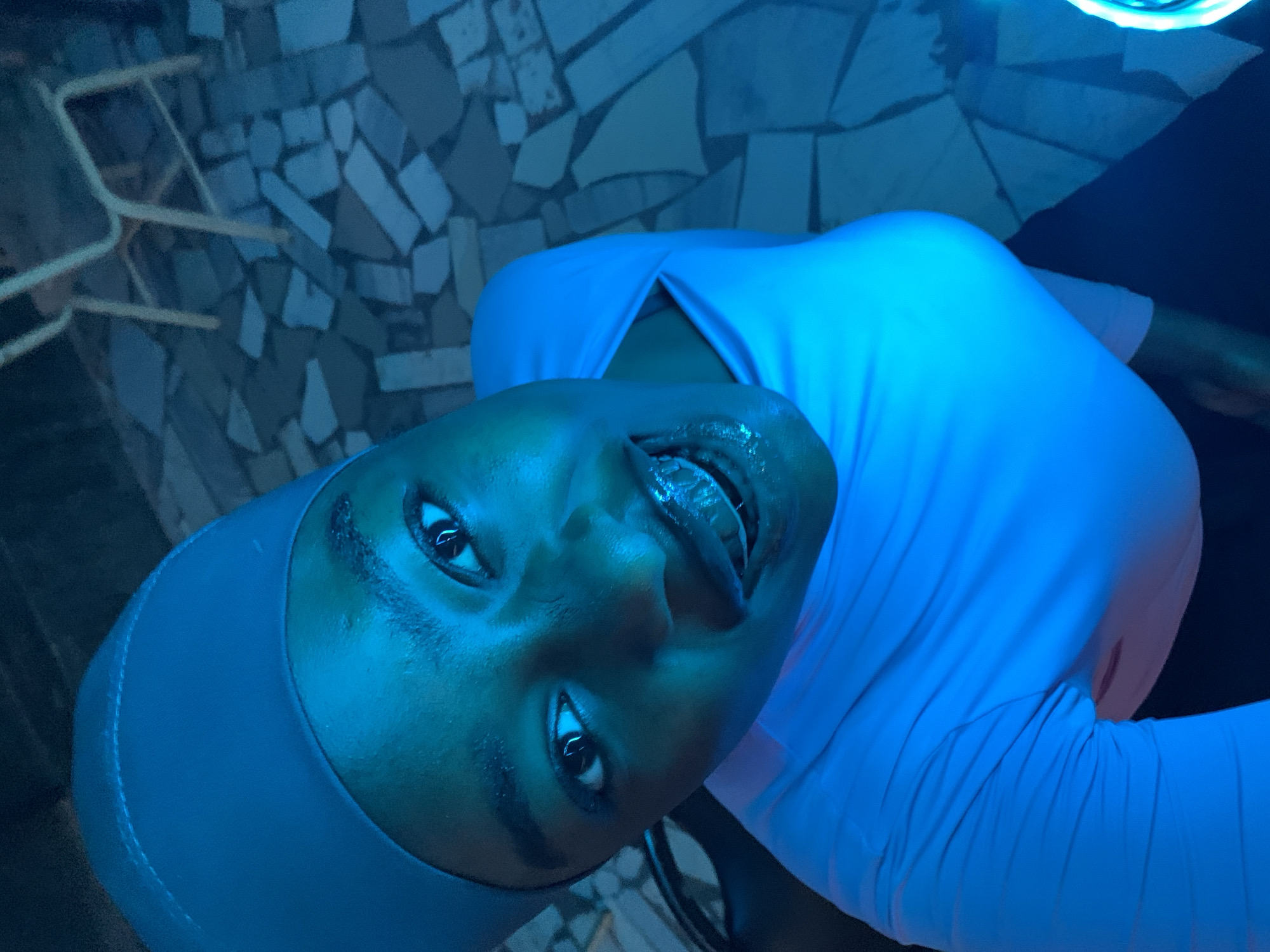 A person wearing a blue cap and a light blue top is looking at the camera. The background appears to be a textured surface, possibly a wall or floor. The scene is bathed in blue light. The image might be taken in a place of New Stadium, Nigeria.