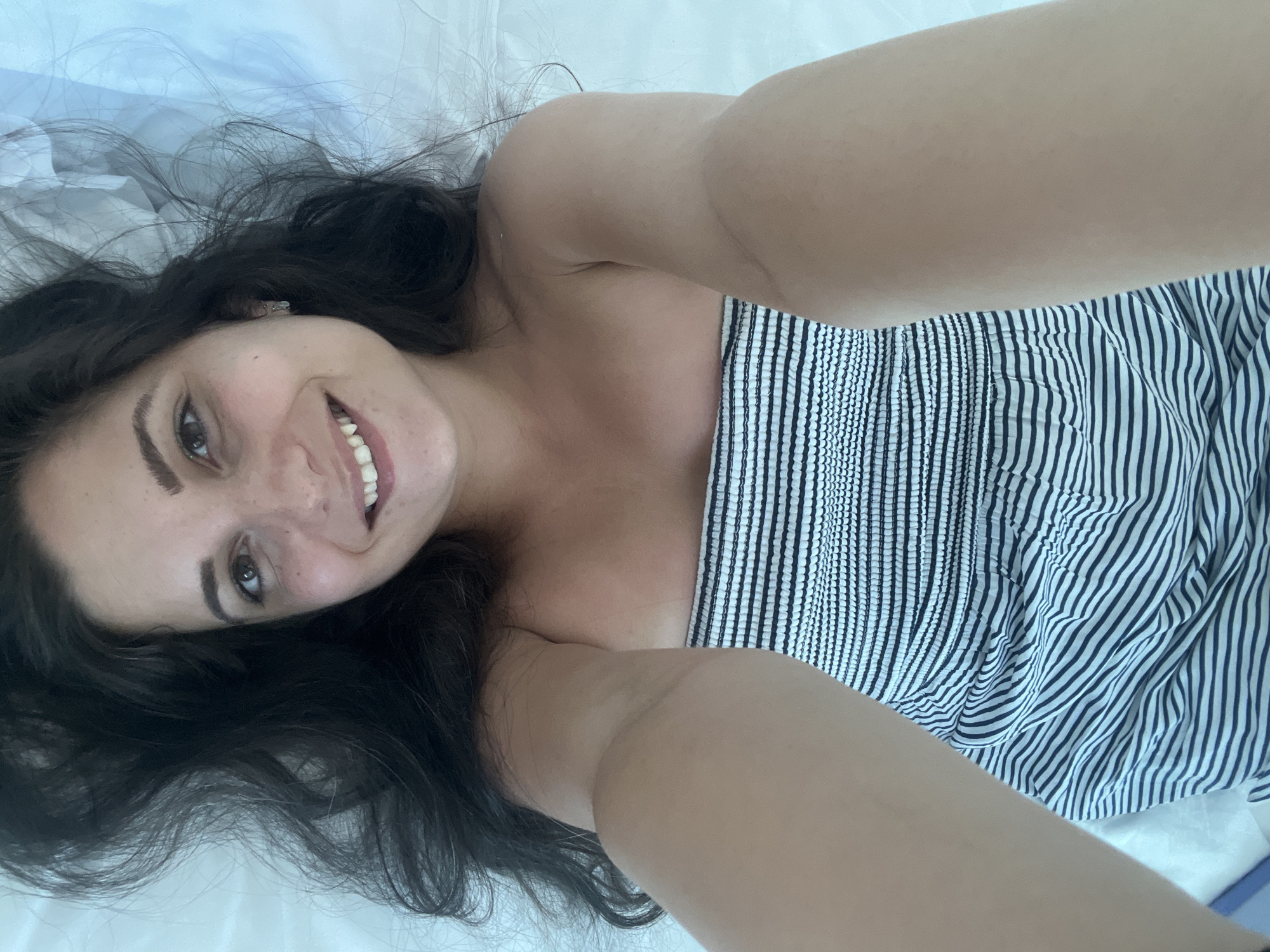 A selfie taken from a first-person perspective shows a woman lying down on a white surface, likely a bed. She has long, dark, wavy hair that spreads out around her head. Her face is turned towards the camera, and she is smiling, revealing white teeth. She has brown eyes and a light dusting of freckles across her nose and cheeks. She is wearing a strapless, horizontally striped dress in black and white. Her arms are extended towards the camera, obscuring parts of her face and torso. The background is a plain white surface, possibly a sheet or a duvet, with some subtle lighting variations suggesting folds or creases. The image appears to be taken indoors, with no visible urban elements to indicate a specific location within Pomorie, Bulgaria.