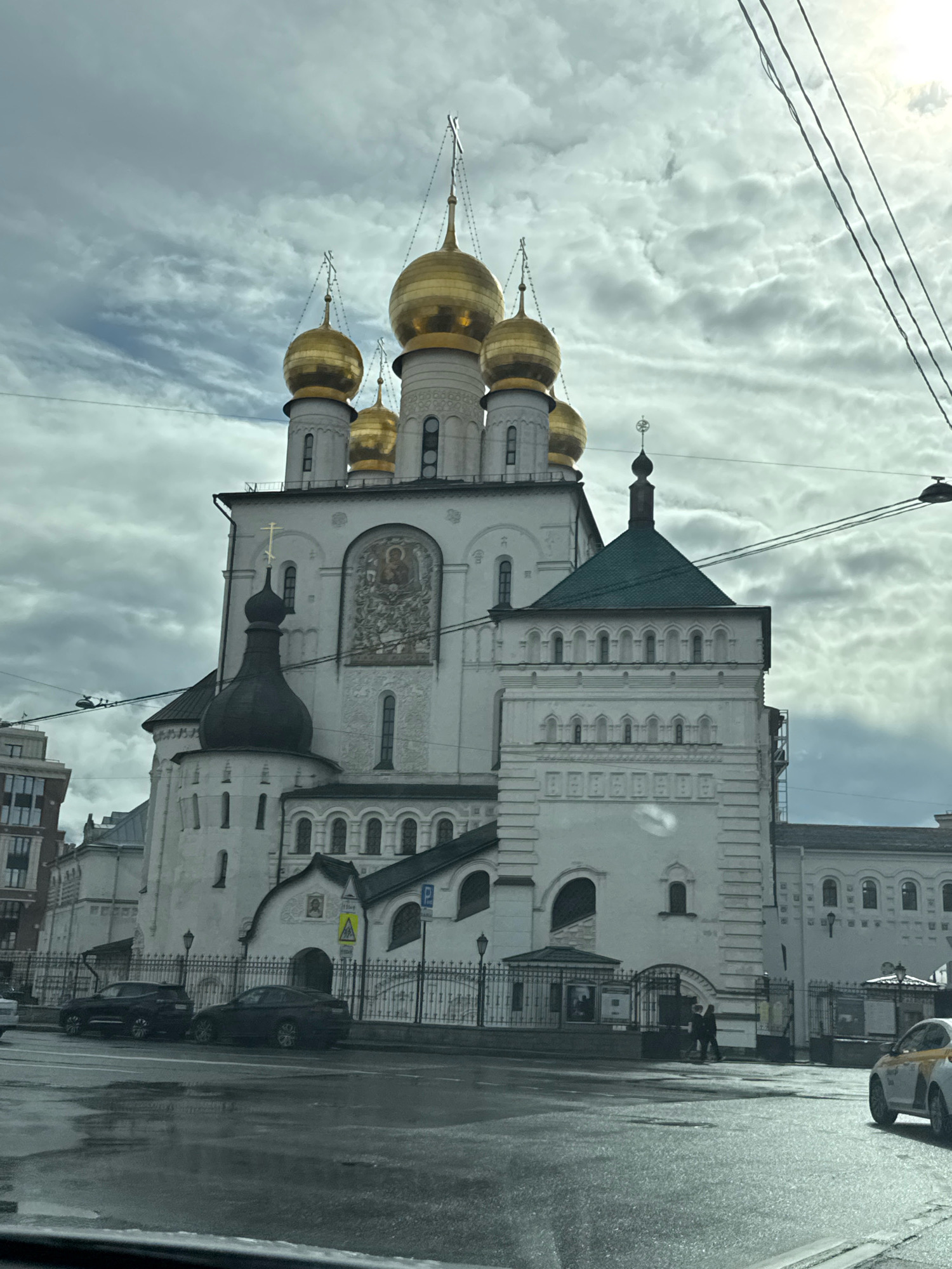 The image shows the Church of the Smolensk Icon of the Mother of God in Saint Petersburg, Russia. It is a white, multi-domed church with golden cupolas. The church is flanked by other buildings and is situated on a street with cars and people. The sky is overcast with clouds.