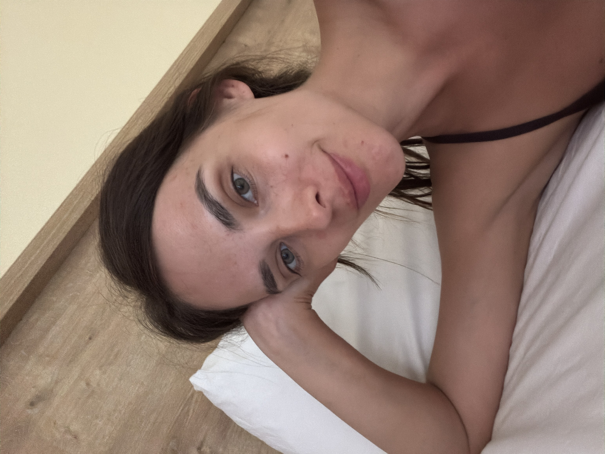 A young woman with dark hair and green eyes is lying in bed, resting her head on her arm. She is looking at the camera with a slight smile. The background shows a wooden headboard and a cream-colored wall. The image appears to be a selfie taken in a bedroom. No specific location in the city of Fethiye, Turkey can be determined from the image.