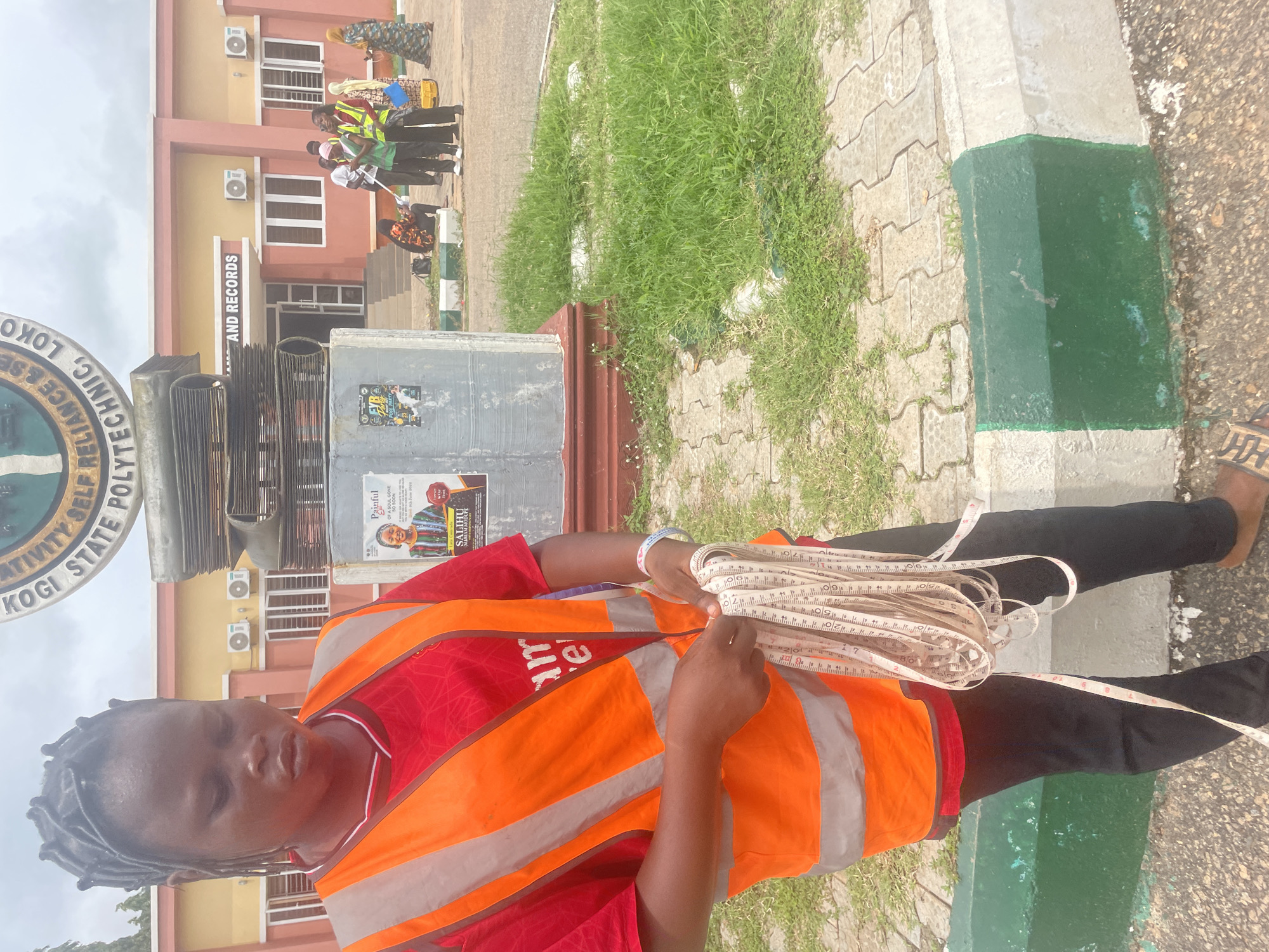 A young woman wearing a red shirt and an orange high-visibility vest is holding a coiled measuring tape. Her hair is styled in small braids. In the background, a building with the inscription "KOGI STATE POLYTECHNIC, LOKOJA" is visible. Several other individuals, some wearing high-visibility vests, are gathered near the building entrance. The area appears to be an outdoor space with paved walkways and grassy areas. The overall setting suggests an educational institution in Lokoja, Nigeria.