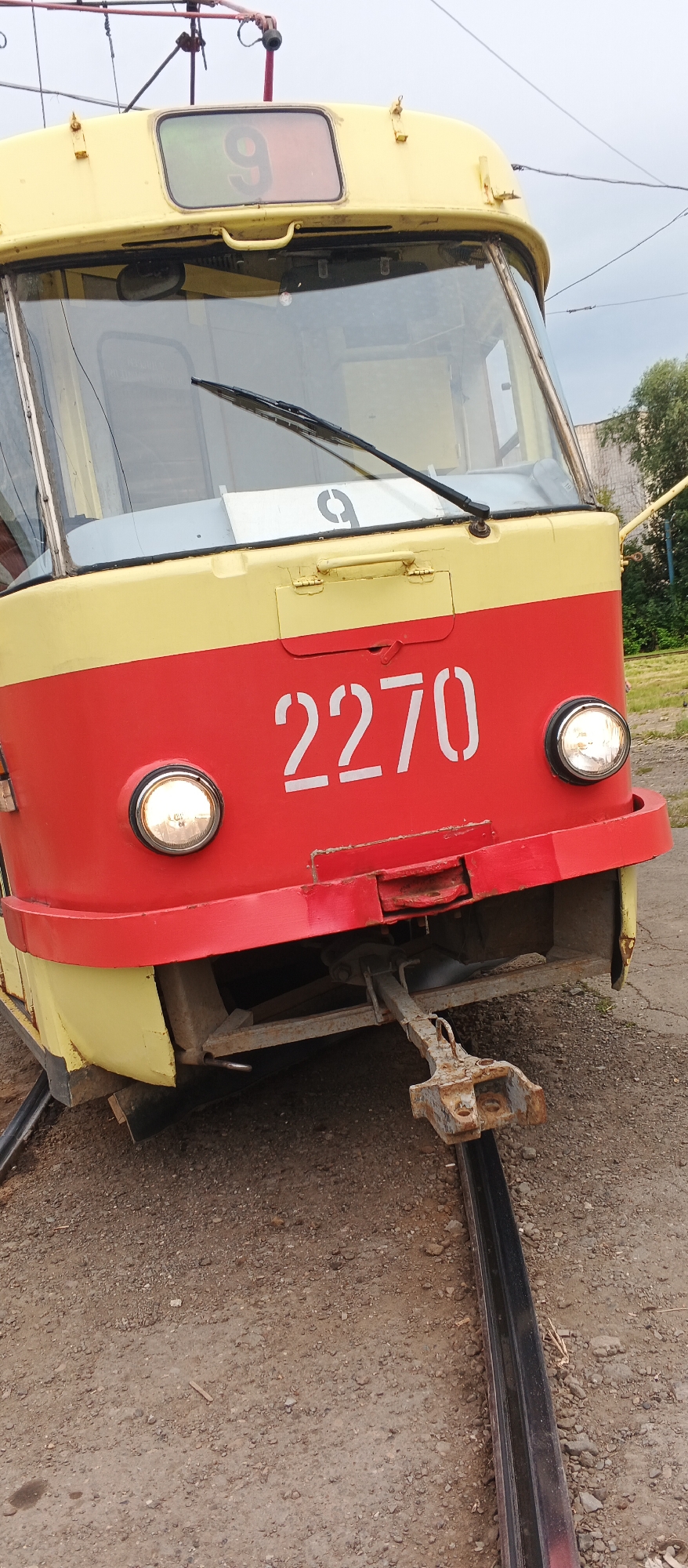 The image shows the front of a yellow and red tram, identified by the number "2270" painted in white on its red lower front panel. Above the windshield, a digital display shows the number "9" within a rectangular frame, indicating it is tram route number 9. The tram is parked on a gravel surface next to a tram track. A metal coupling mechanism is visible extending from the front of the tram, connected to the track. The background shows greenery and buildings, suggesting an outdoor urban or industrial setting. No people are visible in the image.