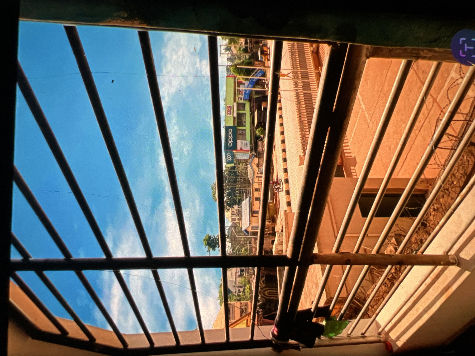 The image depicts a street scene in Lagos, Nigeria, viewed through the bars of a window. The foreground is dominated by dark metal bars, casting shadows across a sunlit balcony area. Beyond the bars, a street is visible, lined with buildings and a light green commercial building with "OPPO" and "VIVO" signage. A person rides a motorcycle on the street, and a yellow motorized tricycle is parked nearby. The sky is bright blue with scattered clouds. The architecture suggests a typical urban environment in Nigeria.