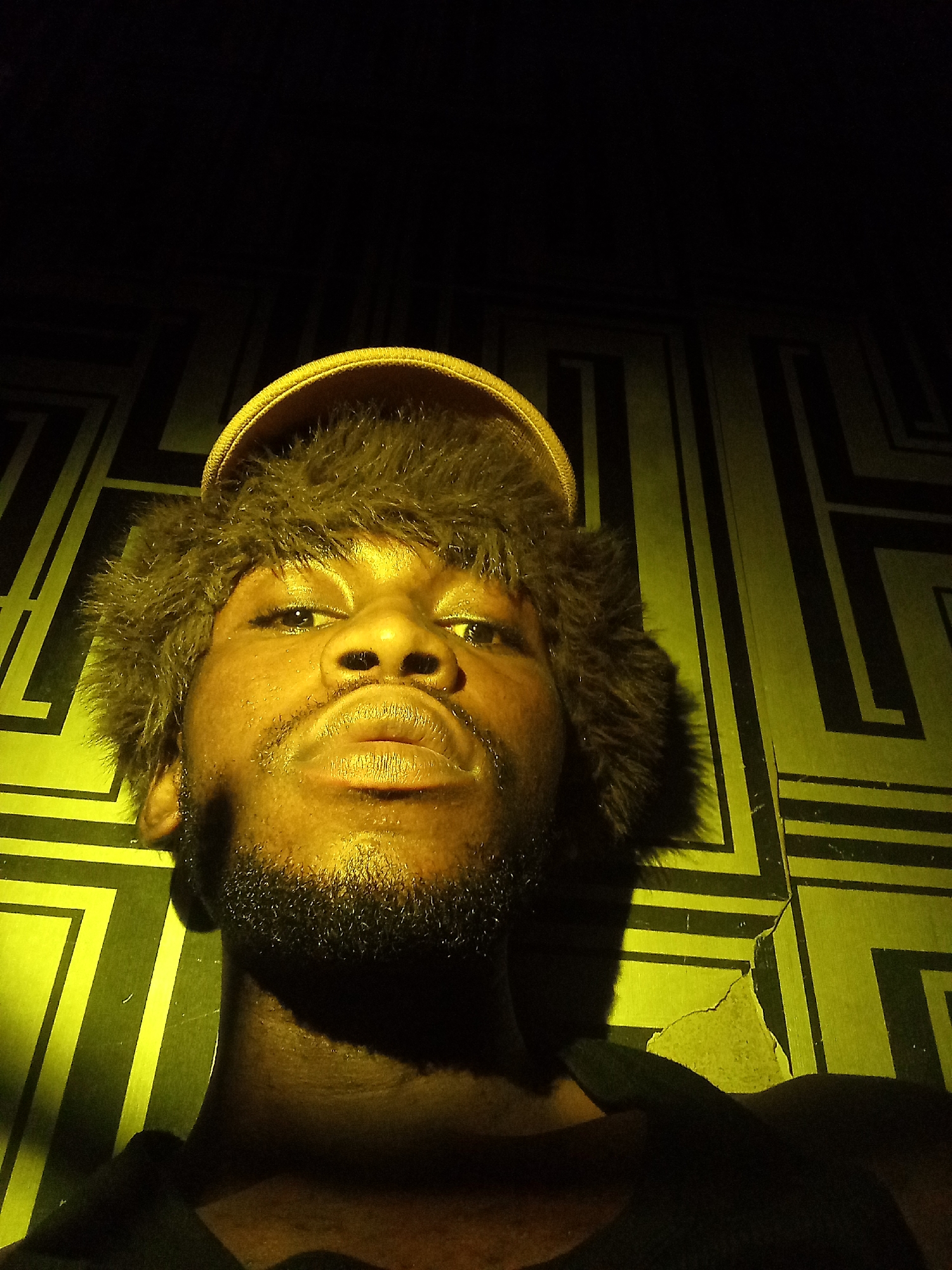 The image is a close-up, low-angle selfie of a Black male wearing a yellow cap with fur trim. He has a light beard and mustache. The background is a wall with a geometric pattern of black and yellow lines. The lighting is dim and casts a yellow hue over the scene. The location is identified as Lokoja, Nigeria.
