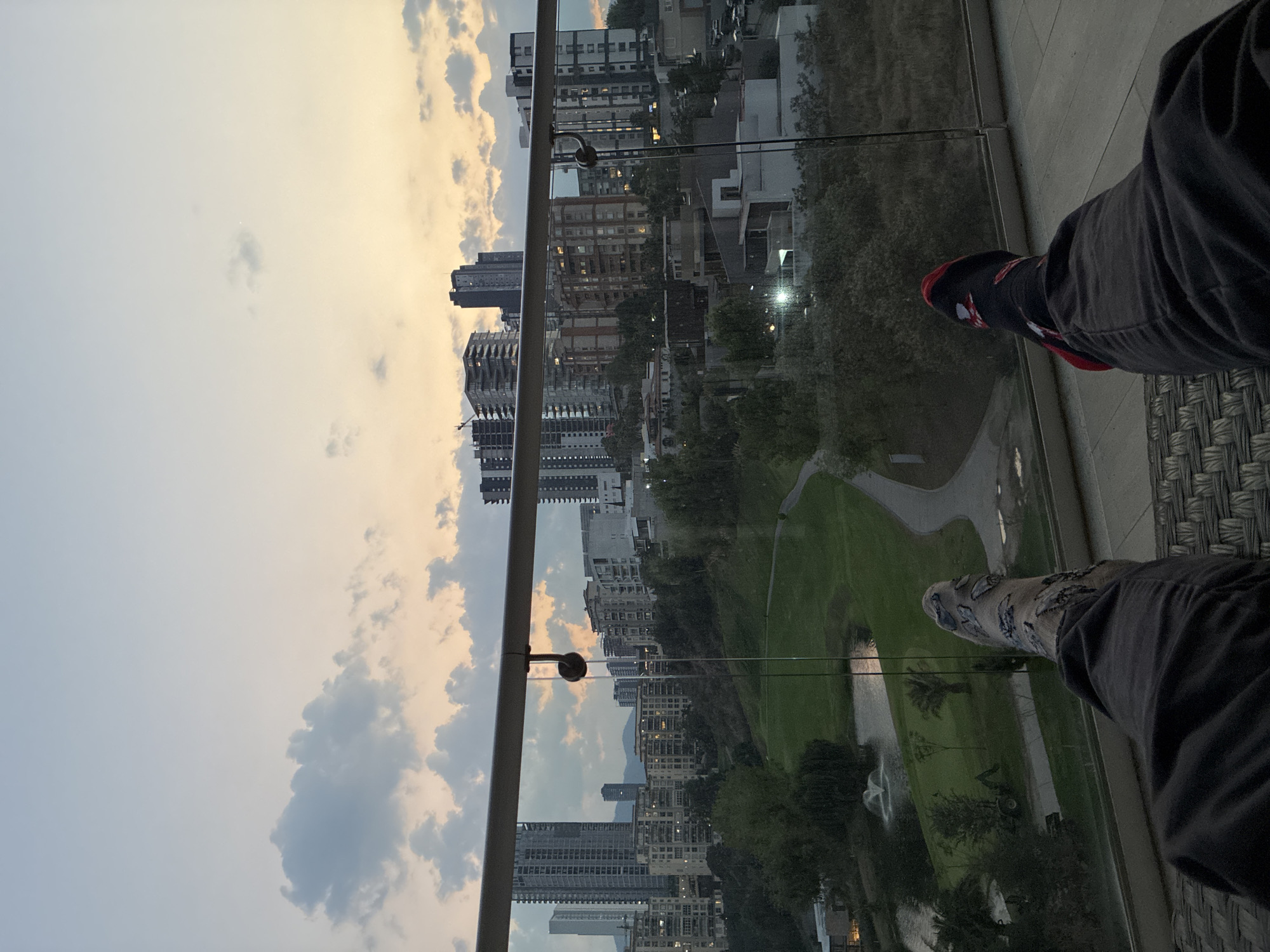 The image is taken from a high vantage point, likely a balcony, overlooking an urban landscape. The foreground shows a partial view of two legs clad in dark-colored pants and different sock designs; one sock is black with red accents, the other light-colored with a darker pattern. A clear glass barrier separates the foreground from the rest of the scene.
Beyond the barrier is a cityscape with numerous tall buildings, interspersed with green spaces. A park-like area with winding paths and a body of water can be seen. The sky is visible above the buildings, showing a mix of light and dark clouds.
The location is identified as Huixquilucan de Degollado, Mexico, based on the urban environment and landscape characteristics.