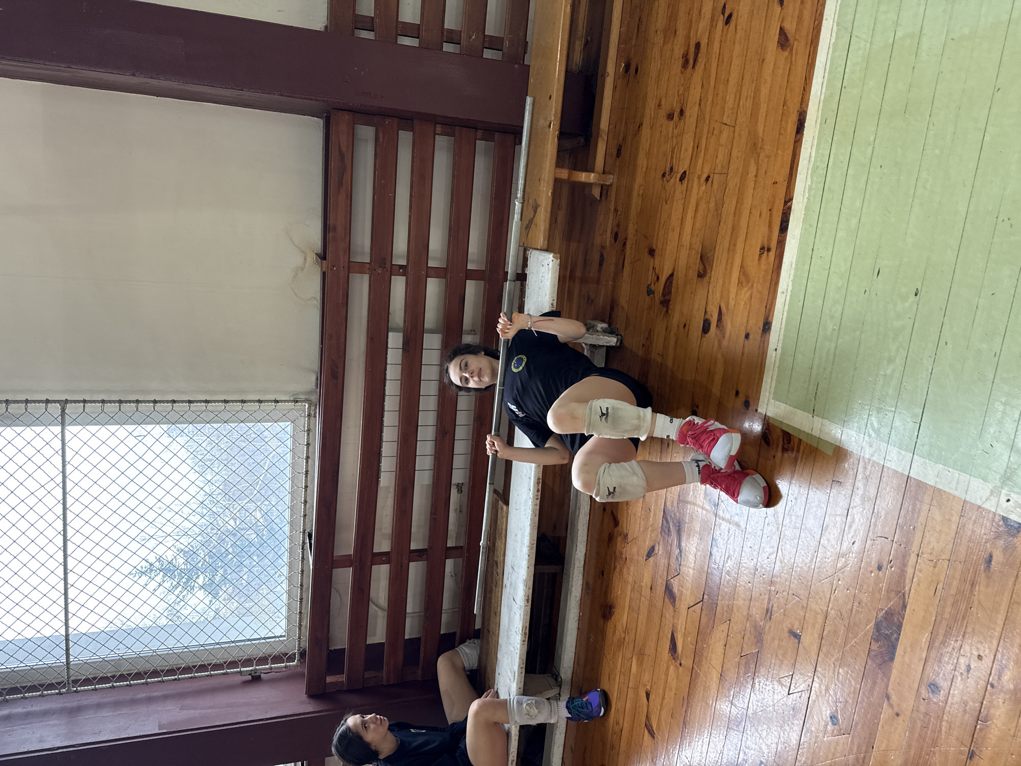 The image depicts a gymnasium or training facility in Sofia, Bulgaria. Two young women are positioned on a wooden floor along a wall, one closer to the camera than the other. Both appear to be athletes, possibly volleyball players, due to their attire which includes dark t-shirts with insignia and knee pads. The woman closer to the camera is also wearing red sneakers. They are both holding onto a metal bar attached to a wooden wall ladder. A window with a wire mesh is visible to the left of the frame. The gymnasium floor consists of wooden planks, partially painted green.