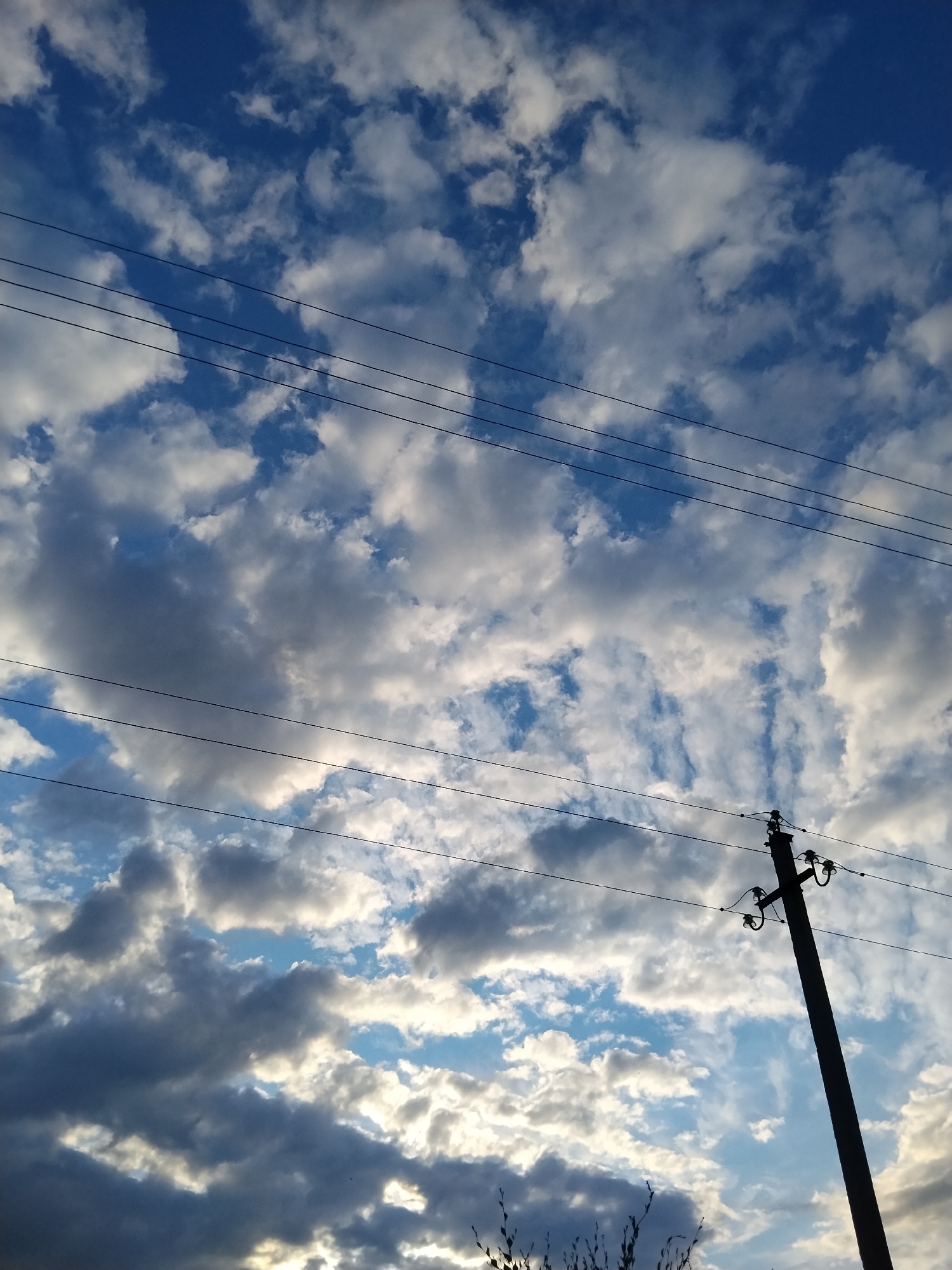 The image is an outdoor, low-angle shot of a sky with scattered clouds and power lines. A single utility pole, silhouetted against the sky, is positioned in the lower right quadrant of the frame. The pole is dark, vertical, and has a crossbar with insulators. Several black, horizontal power lines traverse the sky from left to right across the image. The clouds are a mix of white and gray, with some areas of blue sky visible. The overall lighting suggests it might be late afternoon or early morning. The scene is devoid of people or any other discernible subjects. The image appears to depict a common urban or suburban landscape element.