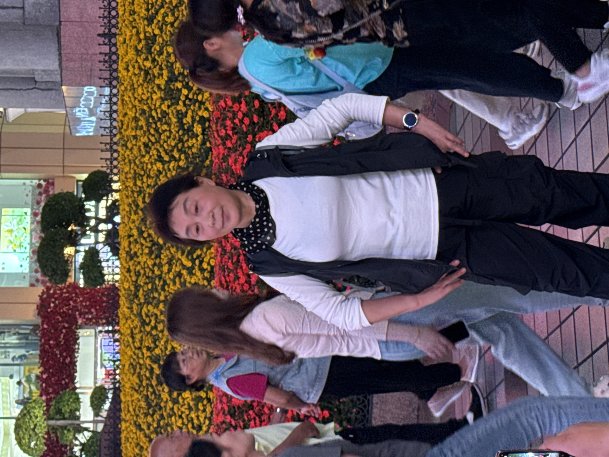 A medium shot captures a group of people, including a woman in a white long-sleeved shirt and dark pants, walking on a pedestrian street in the Yuzhong District of China. The woman is the central focus, looking directly at the camera. To her left is a vibrant flower display in shades of yellow and red. In the background, a glass-fronted building with visible signage and decorative greenery is seen.