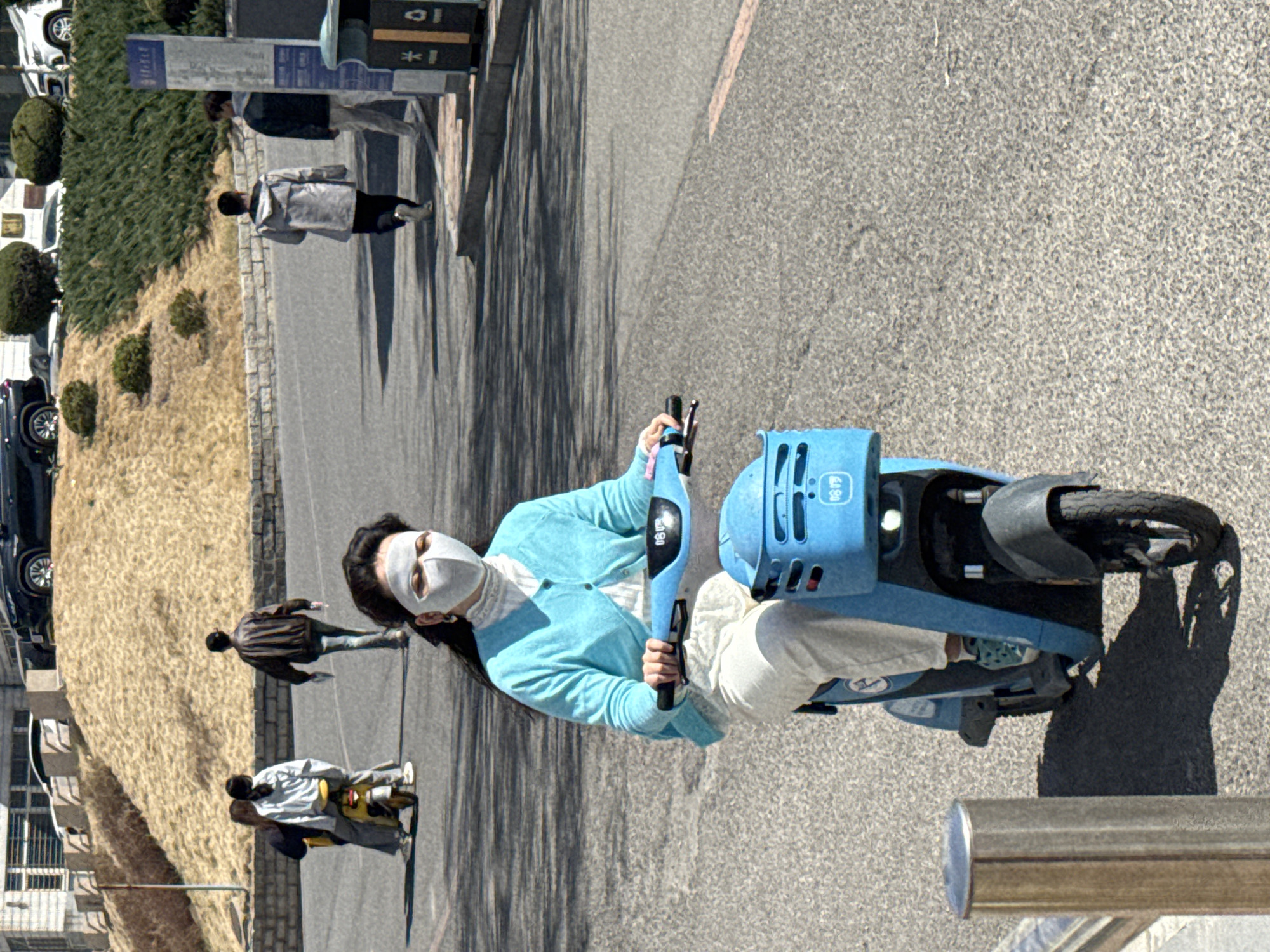 The image shows a woman in Zhongtun, China, wearing a white face mask, a light blue cardigan, and white pants, riding a light blue electric scooter on a paved road. There are pedestrians walking on the sidewalk to the left. The background includes buildings, bushes, and parked cars. There is also street furniture, such as a sign and bollards, next to the road.