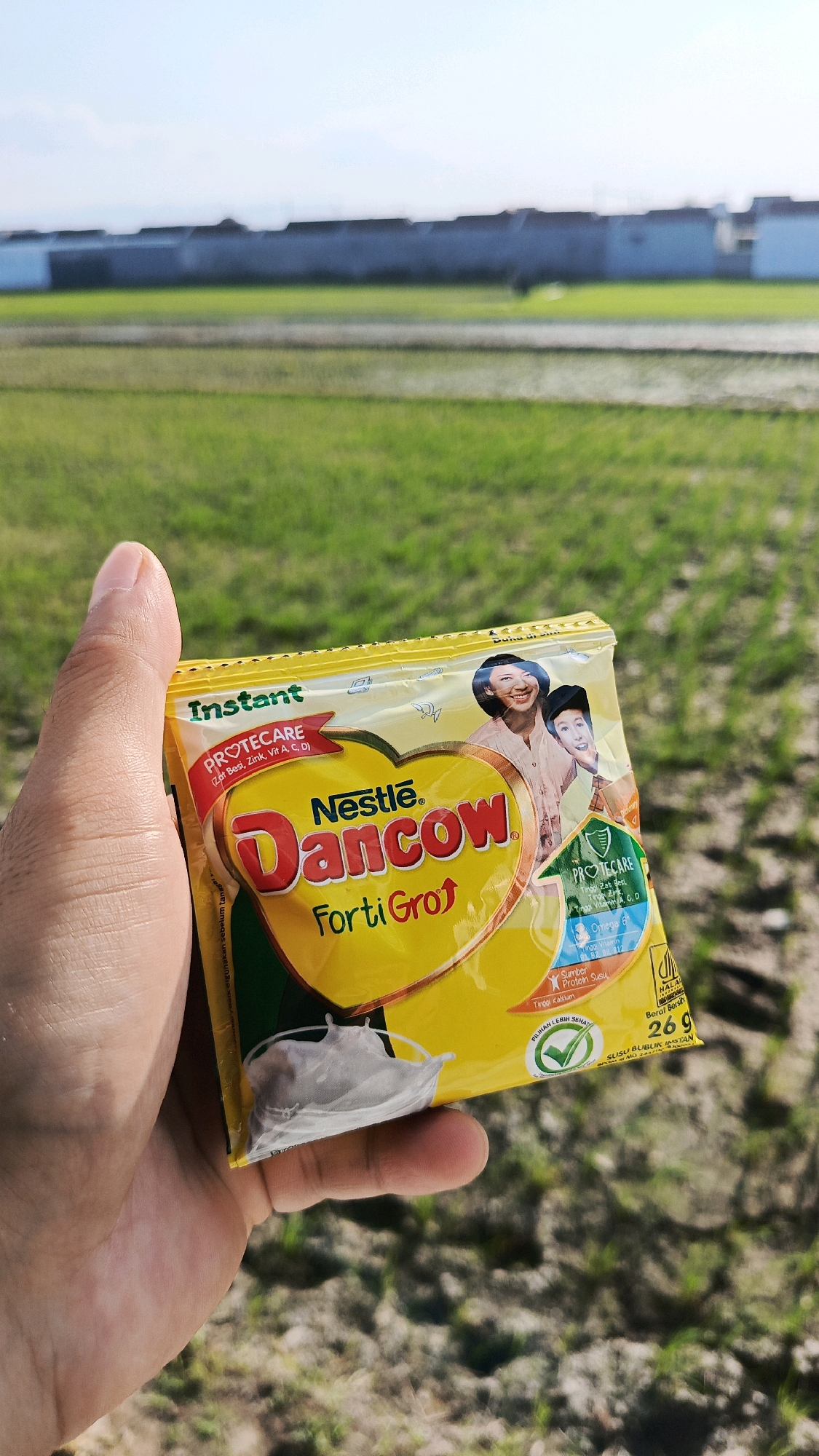 An individual's hand is holding a yellow packet of Nestle Dancow FortiGro instant milk powder. The packet features images of two smiling women and nutritional information. The background shows a bright, sunny day with a clear sky over a green field, possibly a rice paddy, and a row of buildings in the distance. The location is identified as Babadan, Indonesia.