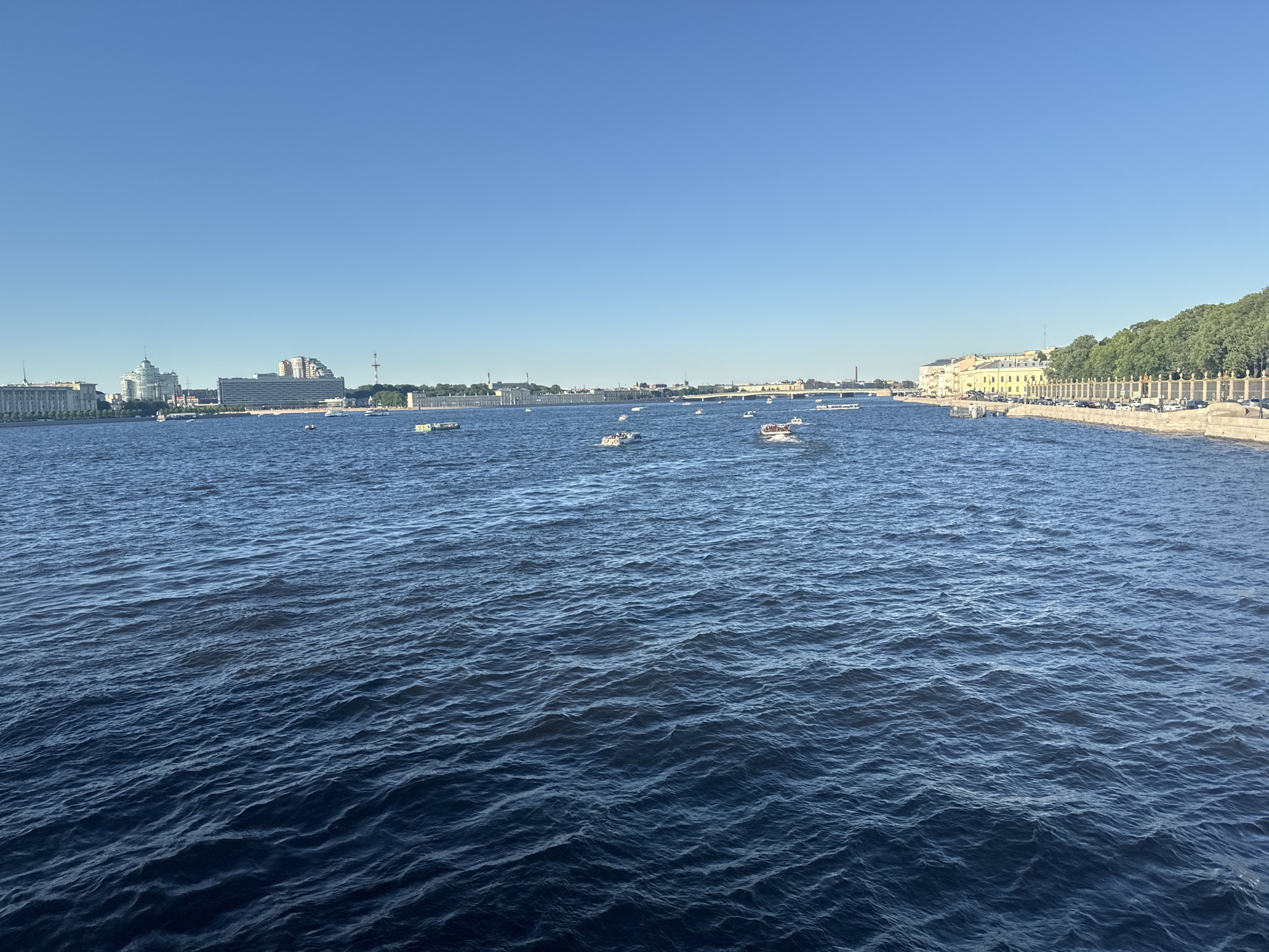 The image shows a wide, blue river in Saint Petersburg, Russia, under a clear, light blue sky. The river is populated by numerous small boats, some carrying passengers. On the left bank of the river, a cluster of buildings is visible, including a prominent domed structure and a modern-looking building with a stepped facade. Further down the left bank, more buildings extend into the distance. The right bank of the river features a long, yellow building with numerous windows, bordered by a decorative fence and trees. A line of parked cars is visible along the embankment. The water surface is rippled, reflecting the clear sky.