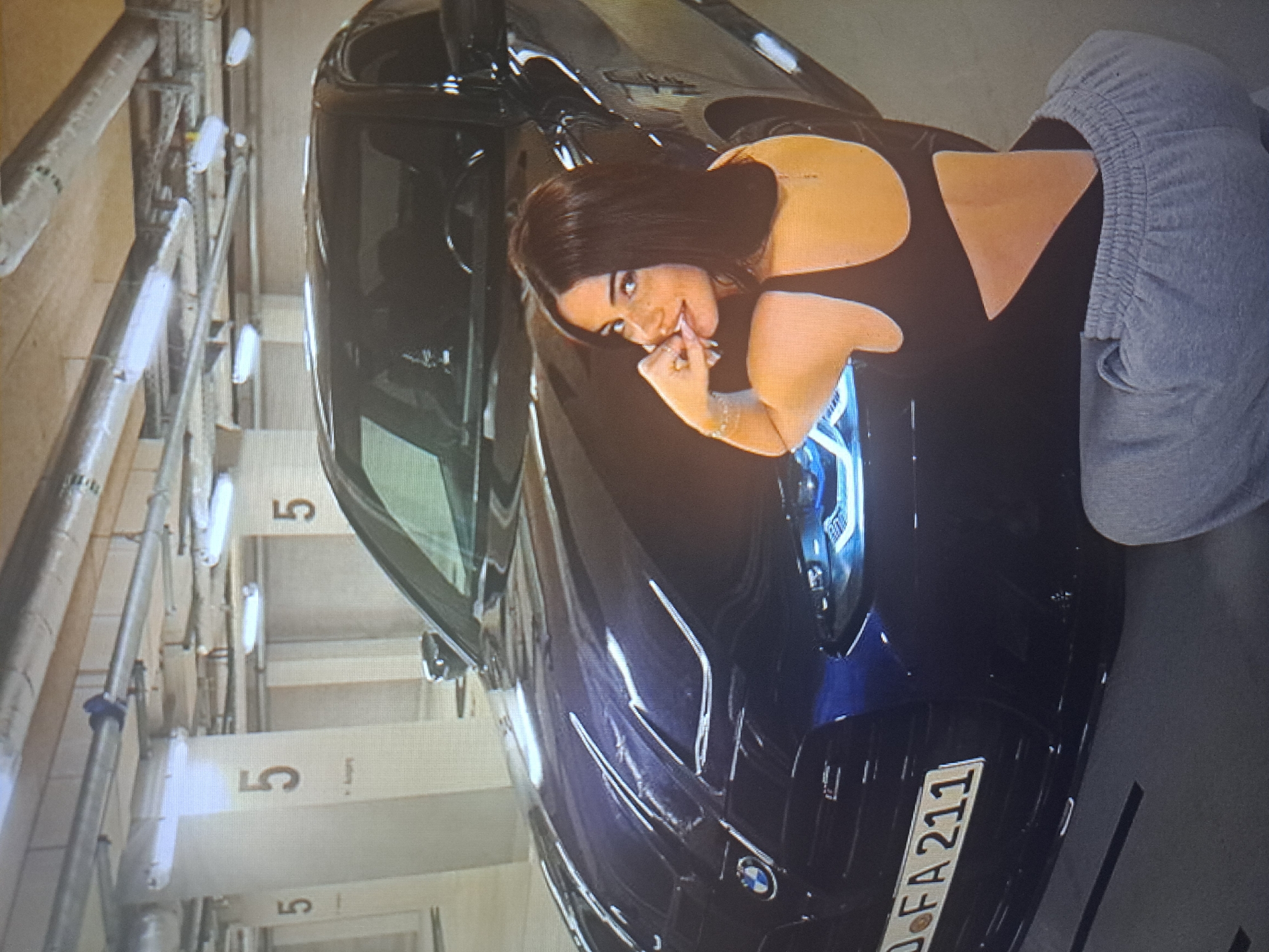 The image depicts a woman posing with a dark-colored BMW in an indoor parking garage. The license plate visible on the car reads "D FA 211". The woman has dark hair and is wearing a black top with a bare back and grey sweatpants. The background of the image shows typical parking garage infrastructure, including pipes and painted parking space numbers, "5". There is no information in the image to confirm that the scene is in Yerevan, Armenia.
