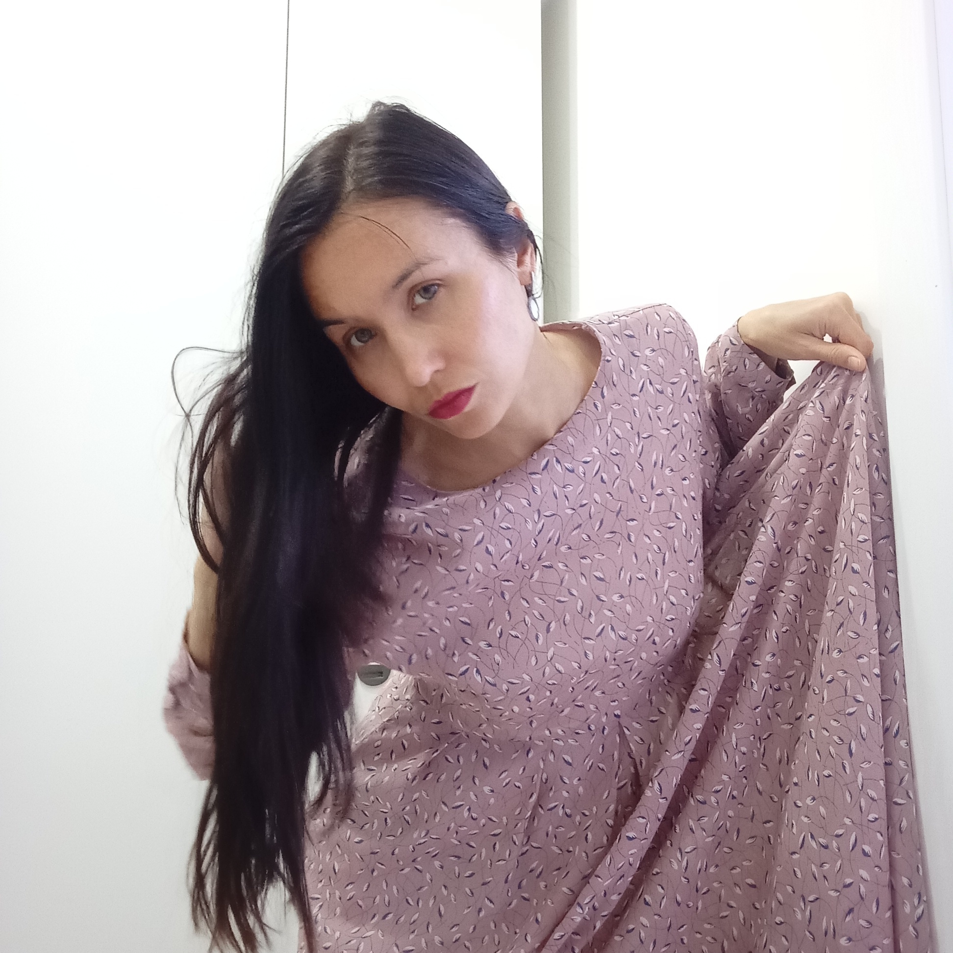 A woman with long black hair, red lipstick, and light skin is standing in front of a white wall or door. She is wearing a long-sleeved, light purple dress with a pattern of small white and purple leaves. She is holding the fabric of her dress up with her right hand, and her left hand is not visible. The background is plain and white. There is no indication in the image that it was taken in Izhevsk, Russia.