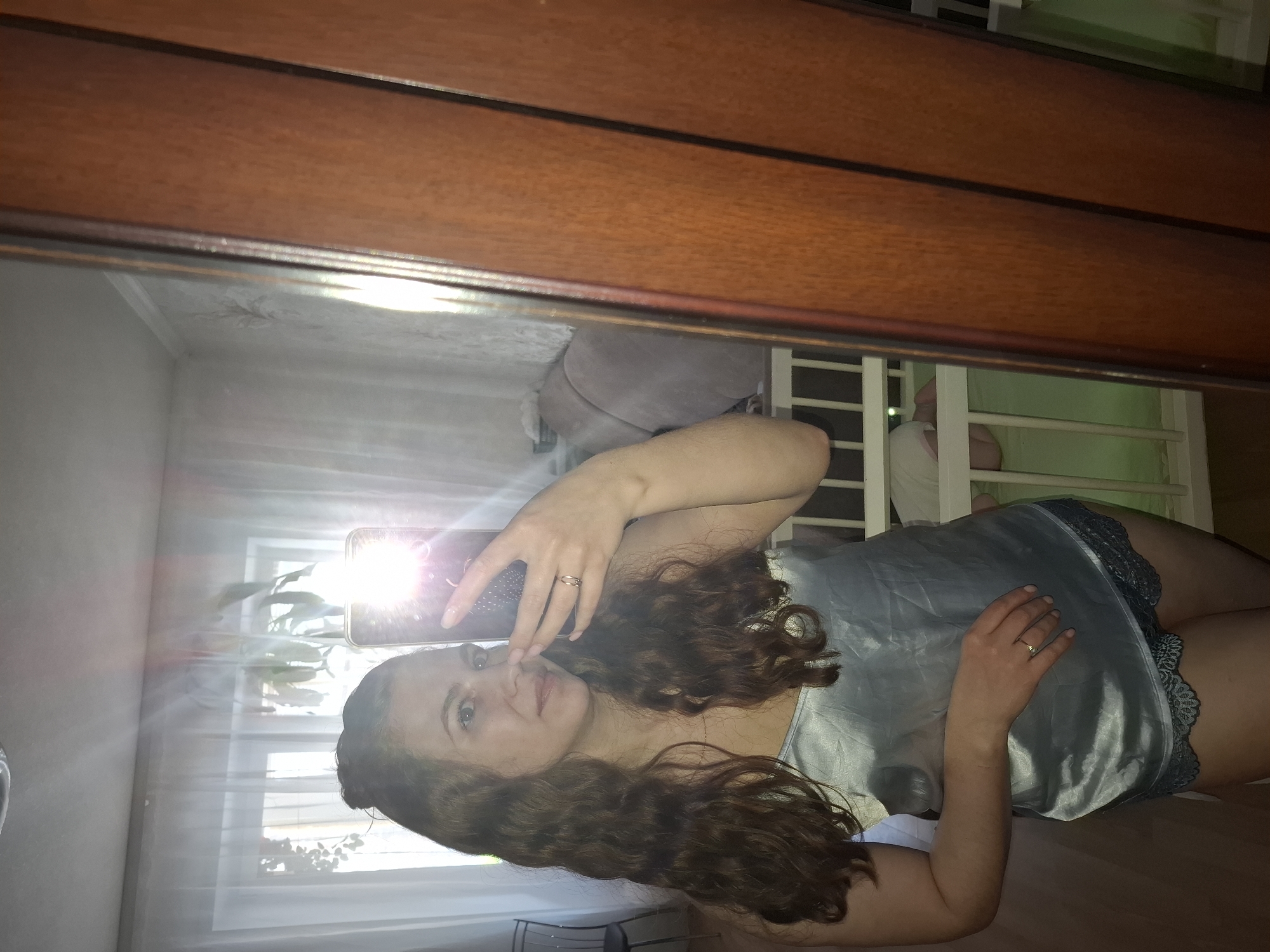 A woman with long curly brown hair and fair skin is taking a selfie in front of a mirror. She is wearing a light blue satin nightgown with lace trim. She is holding a cell phone in her left hand, and there is a bright reflection from the phone's flash. The mirror is framed by a wooden border. The reflection shows a room with white curtains, a potted plant in the window, a couch with grey cushions, and a window with white bars. Based on visual indicators of the room's interior design, the location is presumed to be an apartment or residence. The city of Chaikovsky, Russia is not visually confirmed in the image.