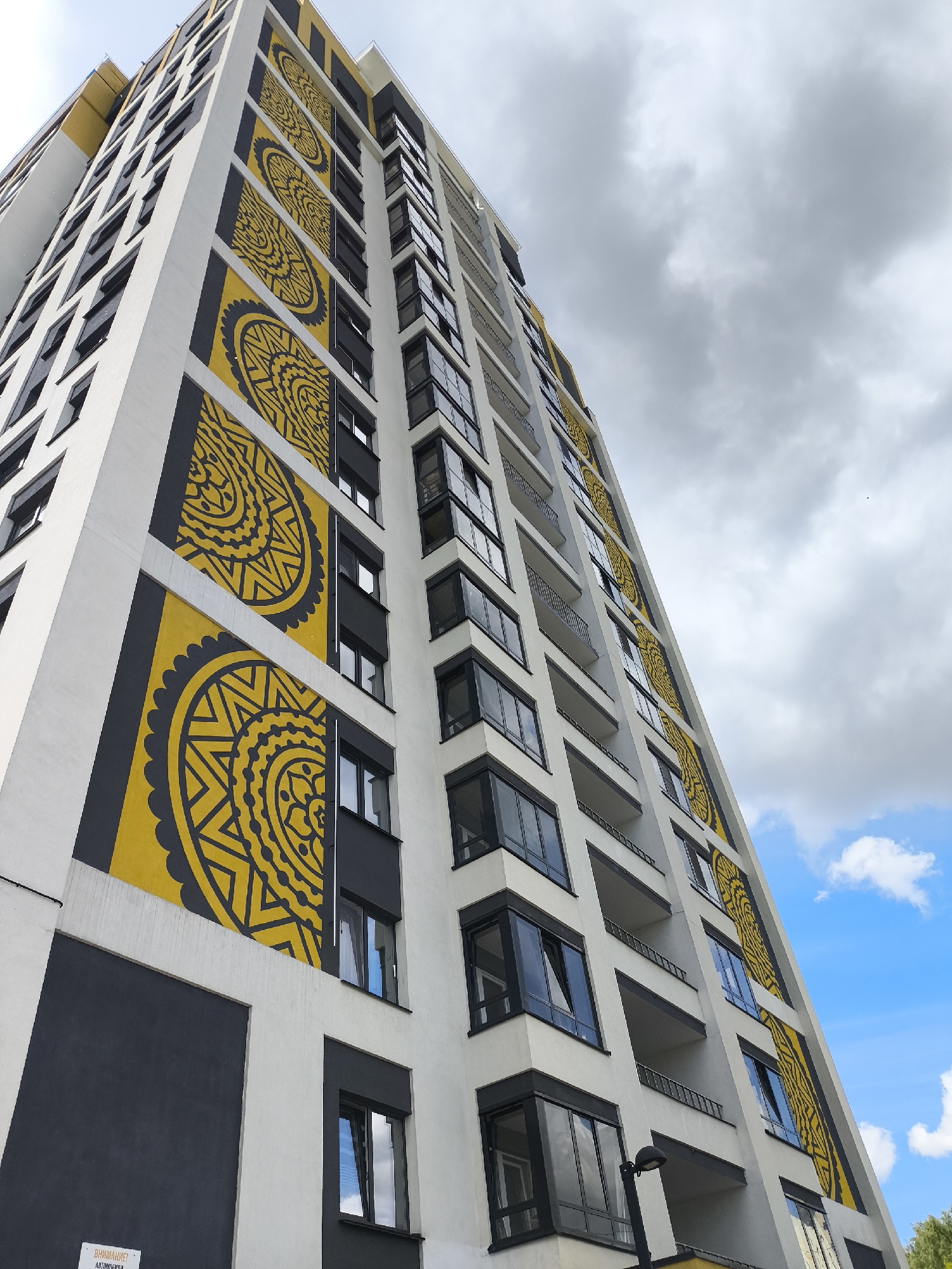 This low-angle, upward-looking photographic image captures a modern, multi-story residential building located in Izhevsk, Russia, under a partly cloudy sky. The building's facade is primarily white and dark grey, accented by prominent vertical sections of vibrant yellow, each featuring a large, intricate dark grey circular geometric pattern resembling a mandala.
The building's design includes recessed dark grey panels and multiple rows of dark-framed windows, some with exterior blinds, and balconies with dark railings. The geometric patterns are consistently displayed in the yellow vertical strips on both visible sides of the building, extending upwards. The patterns are composed of concentric circles and radiating lines, creating a detailed, symmetrical design.
The sky in the upper left portion of the image is overcast with dense grey clouds, transitioning towards the lower right to a clearer blue sky with scattered white cumulus clouds. A black, contemporary street lamp fixture is visible at the bottom right, angled upwards. At the bottom left, a dark grey panel on the building's base features a partially visible white sign with black Cyrillic text, reading "ВНИМАНИЕ!" (ATTENTION!) and "АВТОМОБИЛИ" (AUTOMOBILES), indicating a warning or directive related to vehicles. No individuals or specific actions are depicted in the scene.