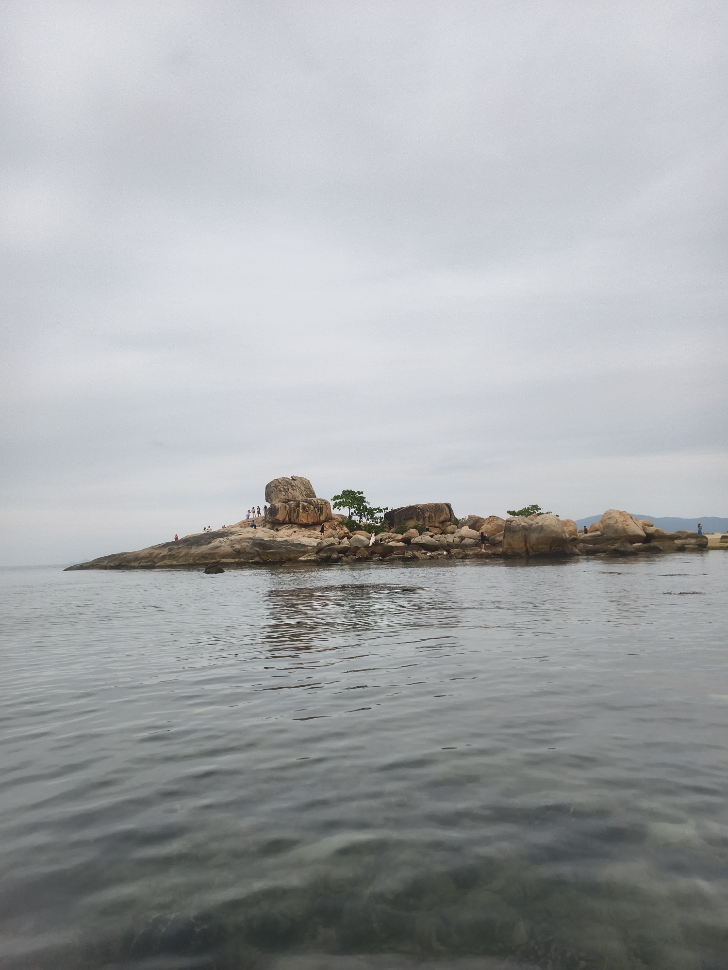 The image shows a rocky island in the ocean under an overcast sky. The water is calm and gray. On the island, there are several large, smooth rocks and some small trees. A group of people can be seen on the rocks, clustered on the left side of the island. The island appears to be located in the city of Bac Nha Trang Ward, Vietnam.