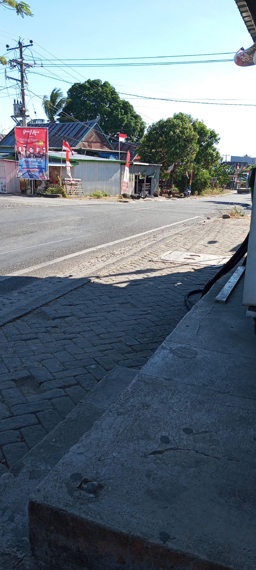 The image depicts a street scene in Pakkanna, Indonesia, on a clear, sunny day. In the foreground, a grey, paved walkway with interlocking bricks runs diagonally from the bottom left to the middle right. A concrete step or ledge is visible on the right side, with shadows cast across the brickwork.

Beyond the walkway is a paved road, with a white line marking the edge. On the opposite side of the road, several structures are visible. A large red banner with text and images of people is displayed on a pole. Next to it, a small, corrugated metal building appears to be a shop, with its entrance open, revealing items displayed inside. In front of the shop, there are several large rocks and a few Indonesian flags, red and white, are planted in the ground and attached to poles. Behind these structures, there are trees and a house with a metal roof. Overhead, electrical wires crisscross the bright blue sky, supported by a utility pole on the left. A motorcycle is parked in front of the shop. Further down the road, other buildings and vegetation are visible, and a vehicle appears to be moving in the distance. The overall impression is of a typical rural or semi-urban street scene in Indonesia, likely a small commercial area or a street with residences and shops.