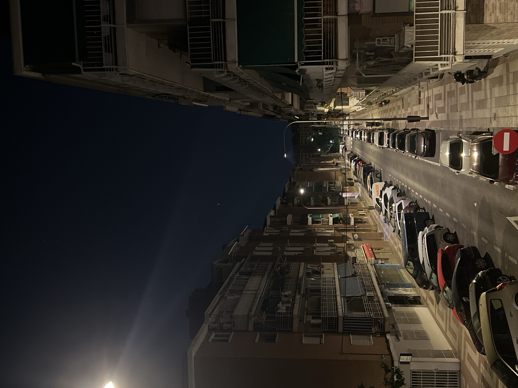 The image depicts a street scene at night in Madrid, Spain. The view is from an elevated perspective, looking down a narrow street lined with tall apartment buildings on both sides. The buildings have numerous windows and balconies. Streetlights illuminate the scene, casting shadows. Cars are parked along the right side of the street. The sky is dark. There are no people visible in the image. The overall atmosphere is quiet and urban.