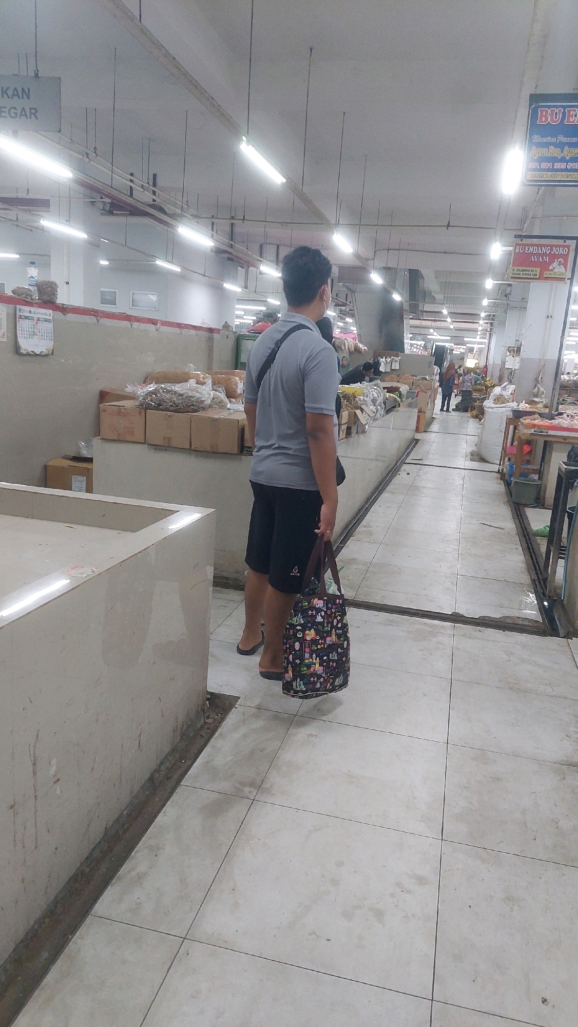 A man stands facing away from the camera in a market in Ponorogo, Indonesia. He is wearing a grey t-shirt, black shorts, and flip-flops. He is carrying a patterned tote bag in his right hand. The market appears to be indoors with a tiled floor and fluorescent lighting. Shelves and stalls filled with goods are visible in the background. Signs in Indonesian are posted on the walls and ceiling.