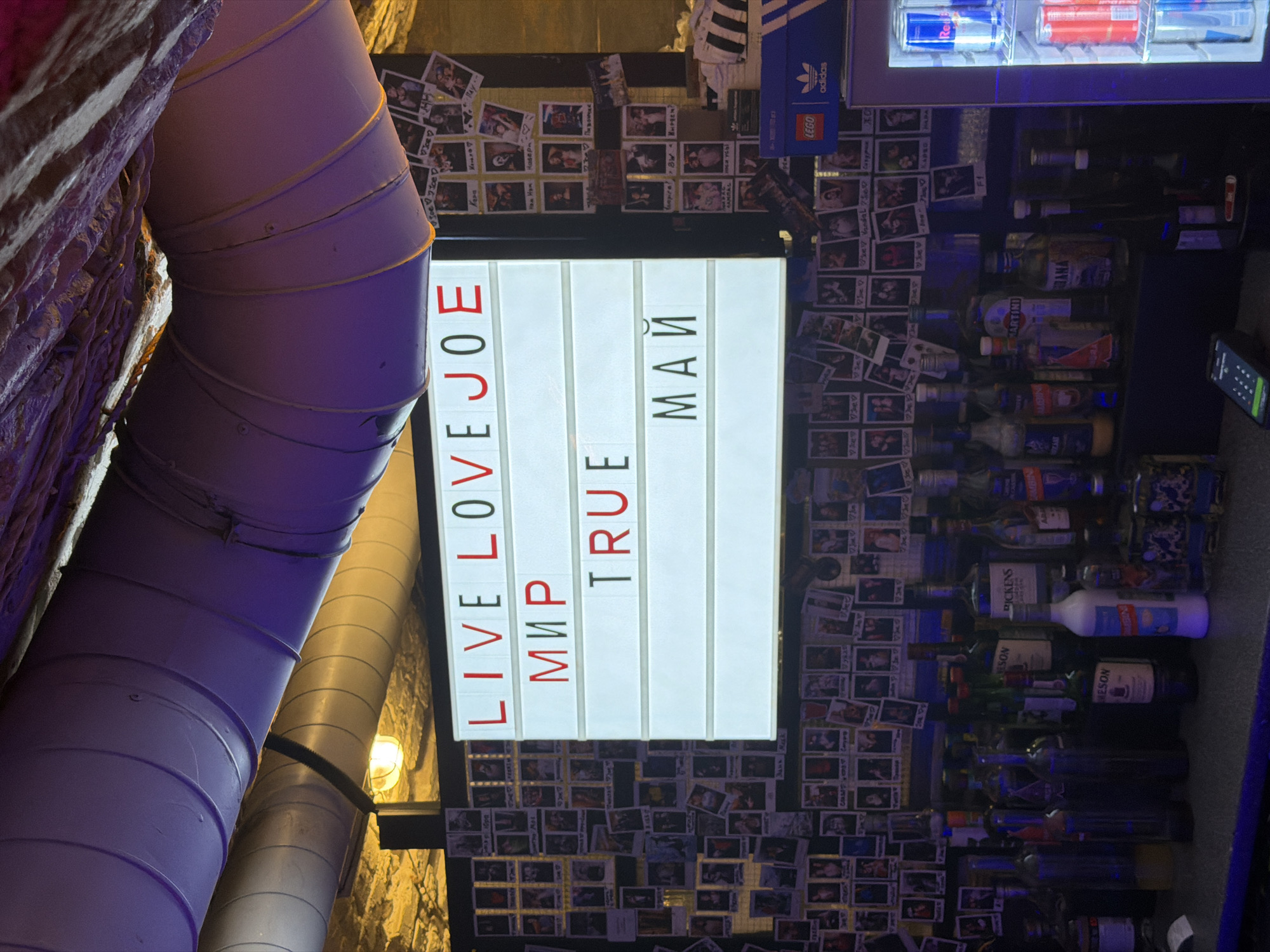 The image captures an indoor scene, likely a bar or entertainment venue in Krasnoyarsk, Russia, characterized by dim, artificial lighting with prominent purple and blue hues. A brightly lit sign board serves as a focal point, displaying a mix of English and Russian text. The English phrases read "LIVE LOVE JOE" and "TRUE," while the Russian words are "МИР" (MIR, meaning 'peace' or 'world') and "МАЙ" (MAY, meaning 'May').
The wall surrounding the sign is densely covered with numerous small, square photographs, possibly Polaroid-style, many featuring people. Various handwritten or printed names are visible on these photos, including "Jann Mesk," "Ye Ye," "beby," "Ham Ham," "Hasep," "Ray," and "V Joe" appearing multiple times.
To the right of the sign, shelves are stocked with an assortment of alcoholic beverages and energy drinks. Recognizable labels include "MARTINI," "PICKENS," and bottles that appear to be "Jameson" (partially visible as "SON" and "MESON"). Several cans of "Red Bull" are also clearly seen. Above these shelves, an "Adidas" logo and a "LEGO" logo are present.
On the left side of the frame, large, curved pipes or ventilation ducts, colored purple, are a prominent feature, extending upwards. These contrast with a rough, textured wall, possibly made of stone or brick, partially visible behind them. Another lighter-colored pipe runs parallel further back. The overall ambiance is eclectic and personal, suggesting a lively social space. The mix of languages on the sign, alongside the given location, indicates a venue with a distinct character.