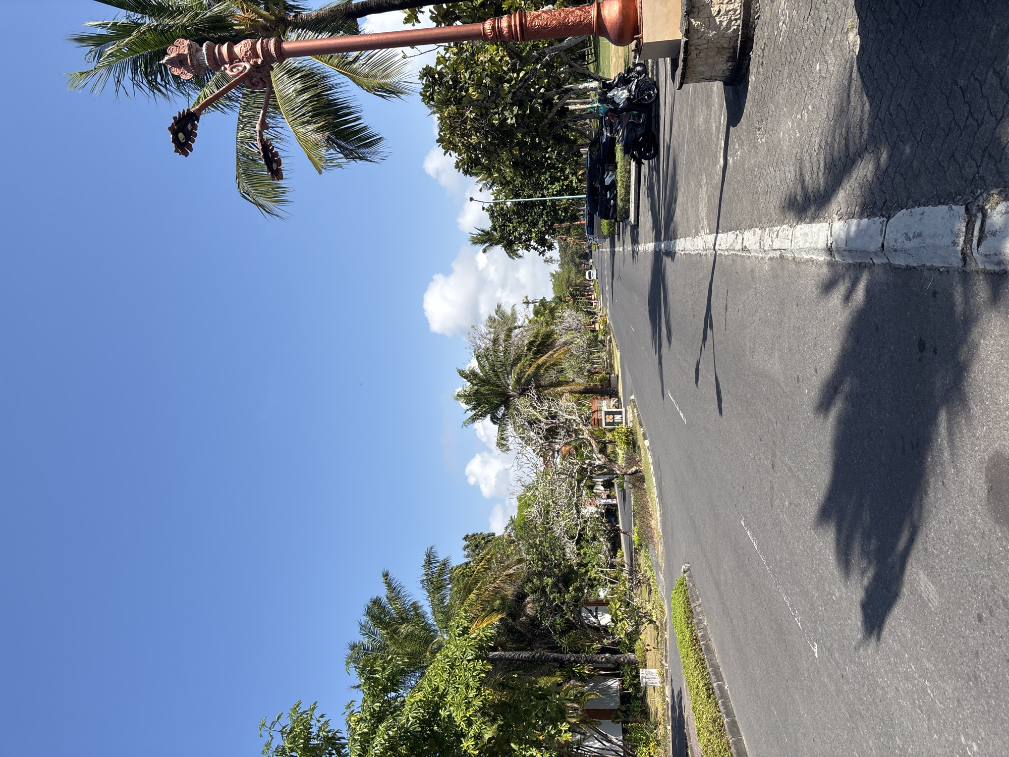 The image depicts a wide asphalt road in Kuta Selatan, Indonesia, flanked by lush green trees and palm trees on both sides. The sky is a clear, vibrant blue with scattered white clouds. On the left side of the road, there are buildings with white walls and tiled roofs, partially obscured by vegetation. On the right side, a sidewalk made of interlocking pavers is visible, along with a decorative lamp post with ornate details and a stone base. Parked on the sidewalk are two black motorbikes and a dark-colored SUV. The road has a white dashed line marking the center and a solid white line bordering the right side. Shadows of palm fronds are cast across the road and sidewalk, indicating a sunny day. A white van is seen in the distance, driving away from the camera.