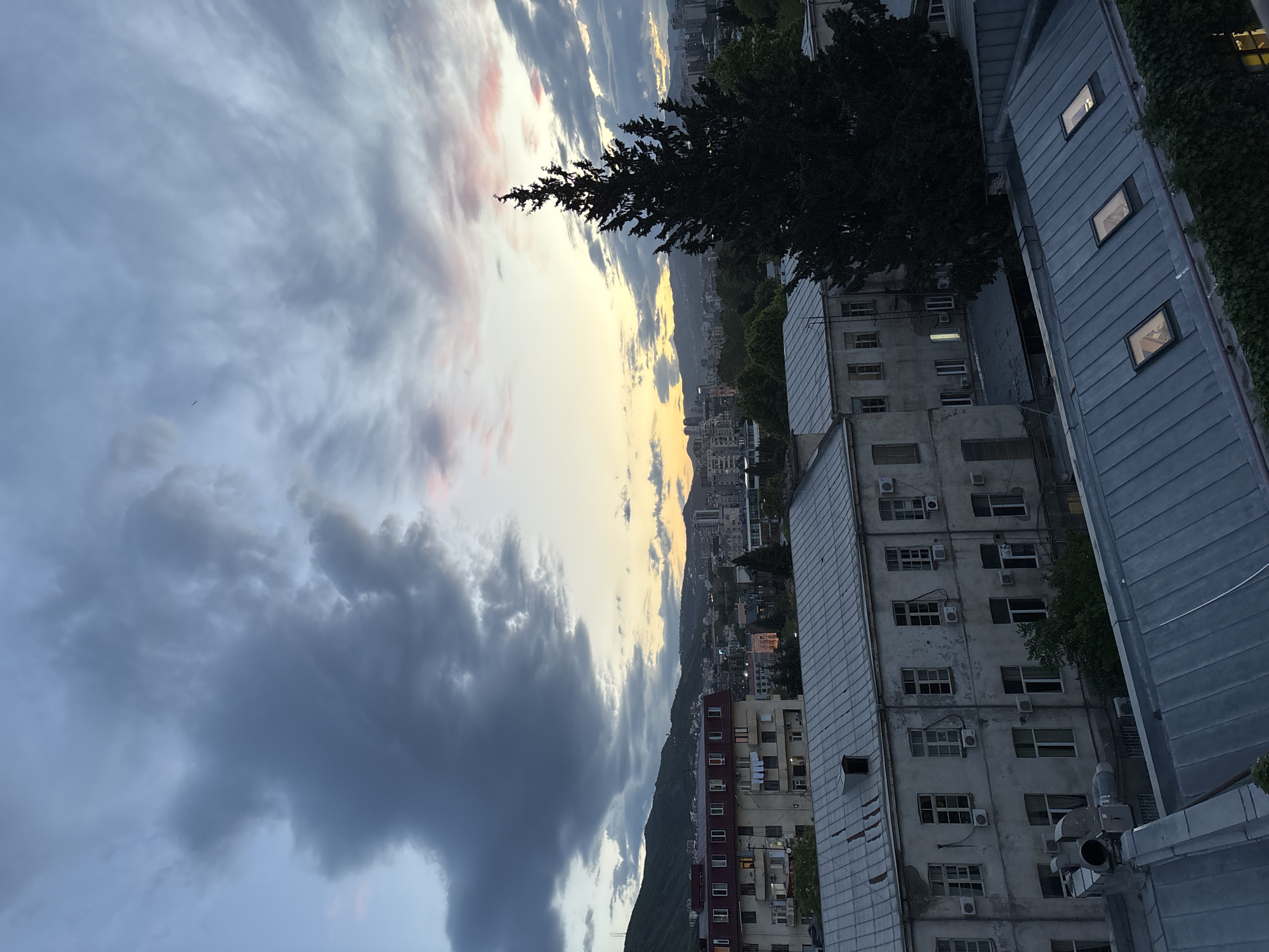 A vertical shot depicts a cityscape at dusk, likely Tbilisi, Georgia, given the architectural style and mountainous background. Buildings of varying heights with Soviet-era architecture fill the frame, some with air conditioning units attached to their exterior walls. A tall evergreen tree partially obscures the view. A mountain range forms a backdrop, with a fading yellow sunset painting the sky. The sky is dominated by dark, heavy storm clouds on the left, contrasting with the lighter, pink-tinged clouds toward the center. The overall lighting suggests a transition from day to night.