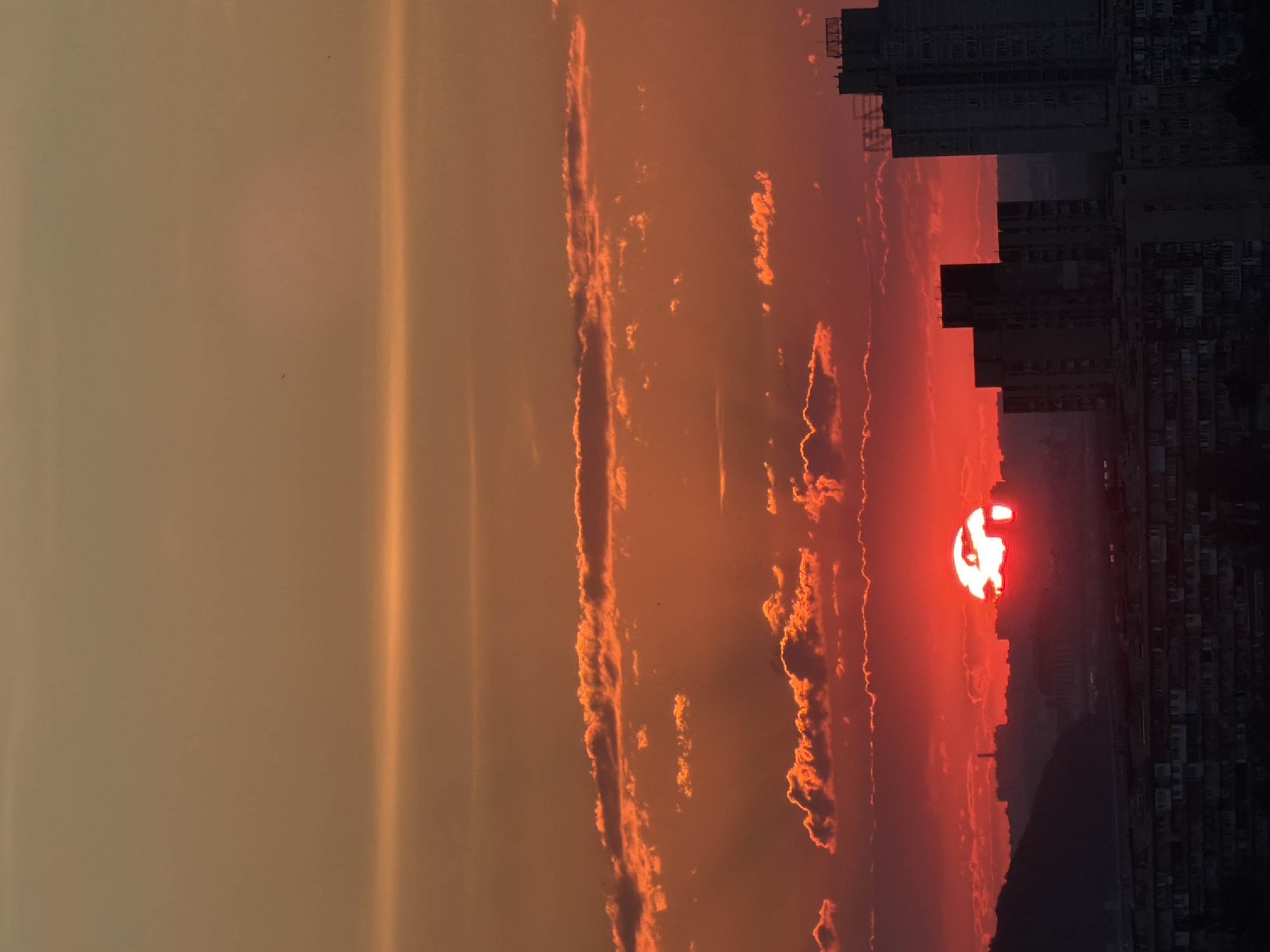 The image captures a sunset over a cityscape, likely Kyiv, Ukraine, given the context. The sun, a vibrant orb of intense red and white light, is partially obscured by the horizon and silhouetted buildings. Wispy clouds, tinged with orange and red hues, stretch across the sky, illuminated by the setting sun. These clouds create a dramatic contrast against the darker elements of the cityscape.
The scene depicts the twilight hours, with the sky transitioning from a warm, hazy orange on the left to a deep red around the sun and a dark, almost black silhouette of buildings on the right. The buildings appear to be apartment complexes or office blocks, tall and densely packed, suggesting an urban environment. A faint hazy glow emanates from the left side of the image, possibly from atmospheric conditions or light pollution.
No people are visible, and the focus is entirely on the natural phenomenon of the sunset and its interaction with the urban landscape. The overall mood conveyed is one of dramatic beauty and urban tranquility as the day concludes. The intense colors of the sky evoke a sense of awe and wonder. There is no visible text in the image.