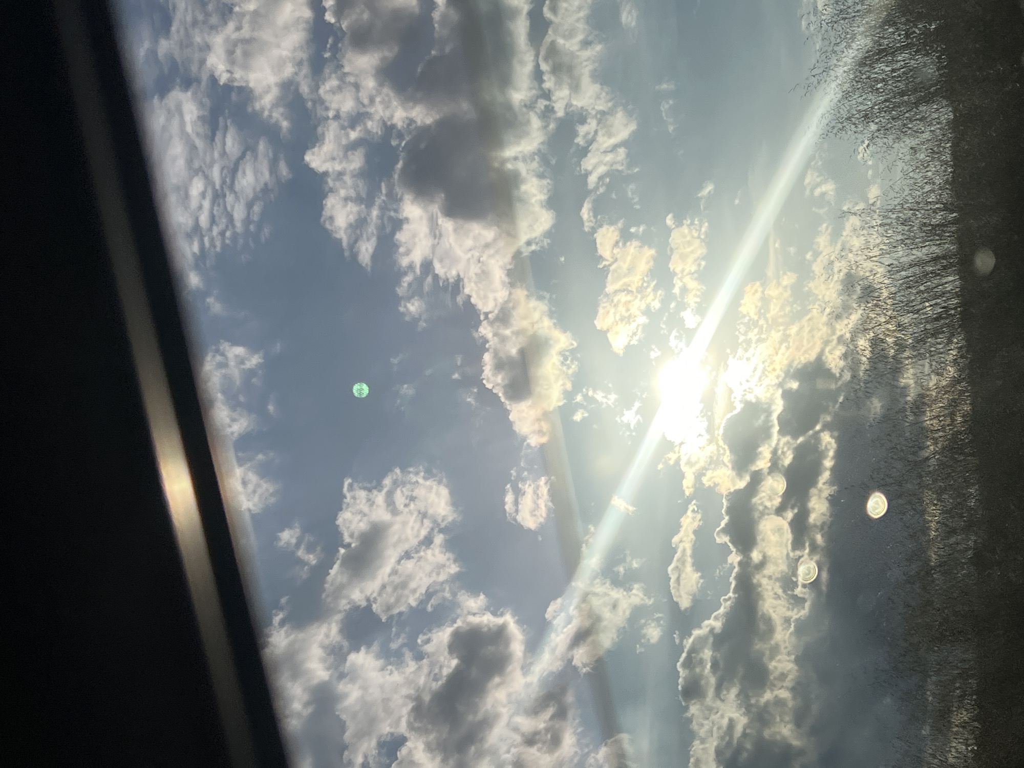 The image displays a bright, partially cloudy sky, seemingly viewed through a window. The sun is a dominant feature in the upper-right portion, casting a strong, diagonal lens flare that stretches across the clouds and blue sky. Numerous cumulus clouds, varying in brightness from brilliant white to shades of grey, are dispersed throughout the frame.
A dark, opaque structure, possibly a vehicle's A-pillar or window frame, occupies the left side of the image. Its inner edge shows a subtle light reflection. On the far right, another dark area with a textured appearance is visible. This texture might be caused by reflections of trees or foliage, or perhaps water droplets or dirt on the outer surface of the glass. Several circular lens flare artifacts are present: a distinct green orb is visible in the middle-left, and two smaller, brighter, concentric ring-like orbs are situated near the center-right, within the textured area.
The scene depicts a sunny daytime environment with a dynamic sky. There is no discernible activity other than observing the sky from an interior perspective. No visible text is present in the image. While the location context is Pesnopoy, Bulgaria, no specific geographical cues are visible within the image itself.