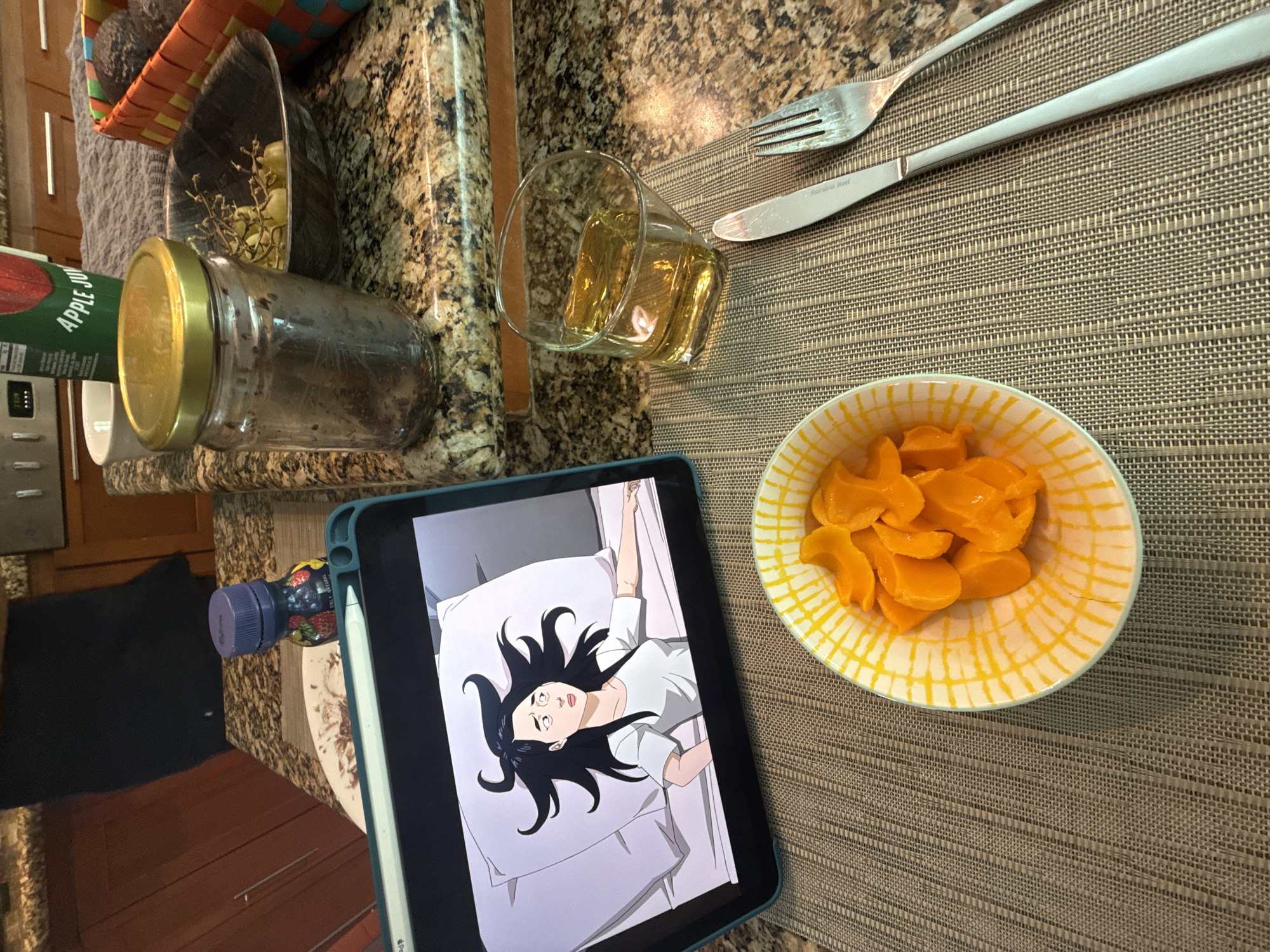 A high-angle, slightly rotated image shows a tablescape arrangement on a brown woven placemat. Dominating the scene are a bowl of orange fruit slices, a tablet displaying an animation of a person lying on a bed, and silverware (fork and knife). To the left, there's a jar with a gold lid, a glass with a light-colored liquid, and a small bottle with a purple lid. The surface beneath this arrangement is a speckled brown countertop. The location of San Bernardino Tlaxcalancingo, Mexico cannot be verified within the content of the image.