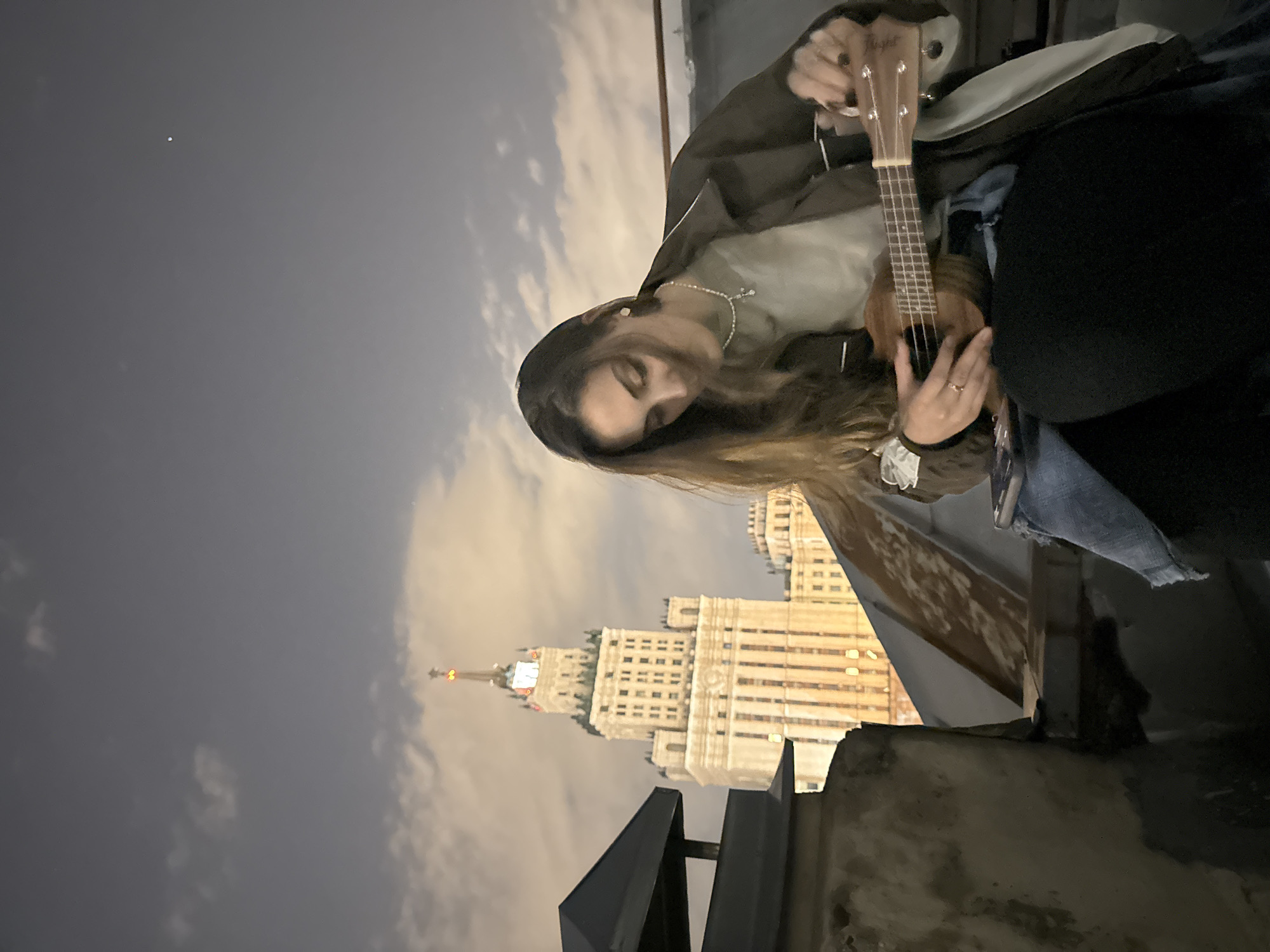 A woman is sitting on a rooftop playing a ukulele at night. The Seven Sisters building (Kudrinskaya Square Building) in Moscow, Russia is visible in the background. The woman has long brown hair and is wearing a brown jacket and a necklace. The sky is cloudy.