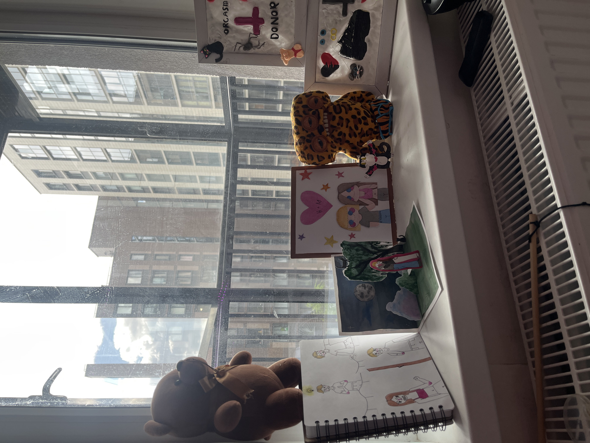 The image shows a windowsill inside a building, with a view of other buildings across the street. The windowsill is cluttered with various objects, including a teddy bear, drawings, framed artwork, and a stuffed leopard. The window is dirty, obscuring the view of the city. The city is not Saint Petersburg, Russia. A radiator is visible on the right side of the image.