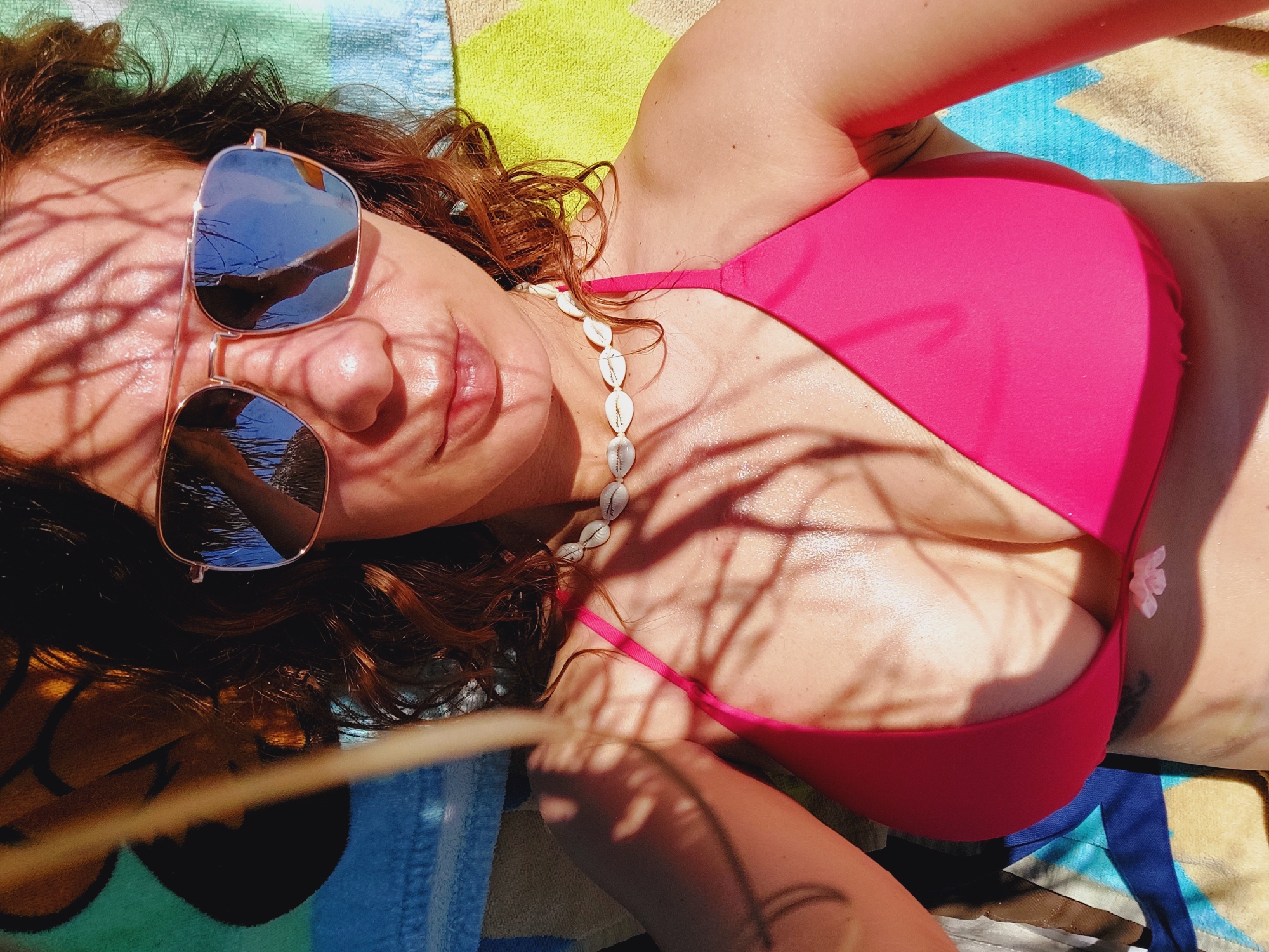 A woman in a pink bikini top and sunglasses lies on a colorful striped beach towel, presumably in Sozopol, Bulgaria. Sunlight filters through palm fronds, casting dappled shadows on her face and chest. She wears a white shell necklace. Her left arm is visible, resting outside the frame. The image is a close-up, focusing on her upper body.