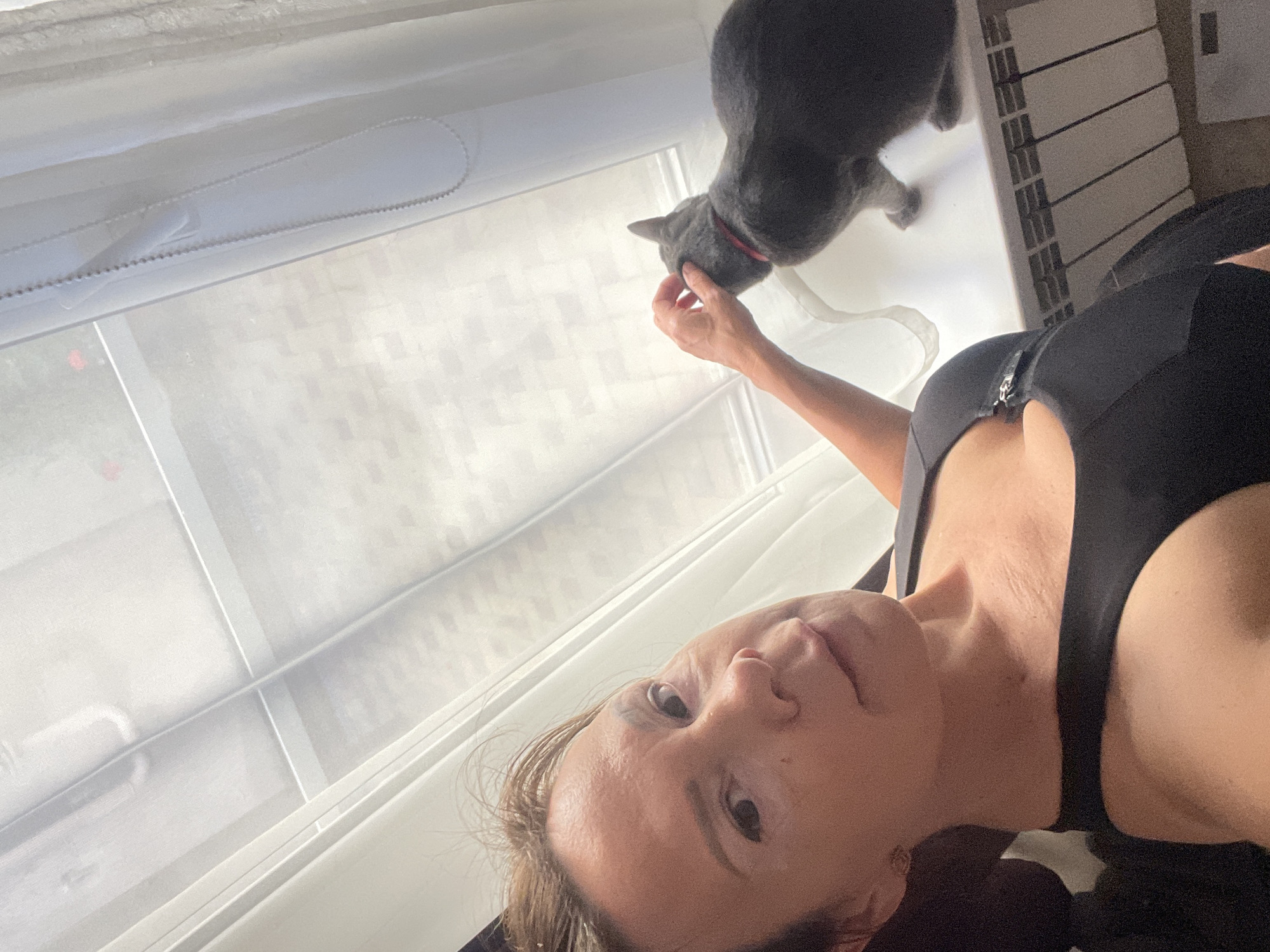 In this selfie, a woman is looking towards the camera with a relaxed expression. She is wearing a black tank top with a zipper detail at the front. To her left, a gray cat with a red collar is leaning in, appearing to interact with her hand. The background suggests an indoor setting near a window with sheer white blinds. The lighting indicates it could be daytime. The image has a candid and intimate feel, possibly capturing a moment of playfulness between the woman and her pet. There is no visible text in the image. The location context of Sochi, Russia, does not provide any specific visual cues in this image.