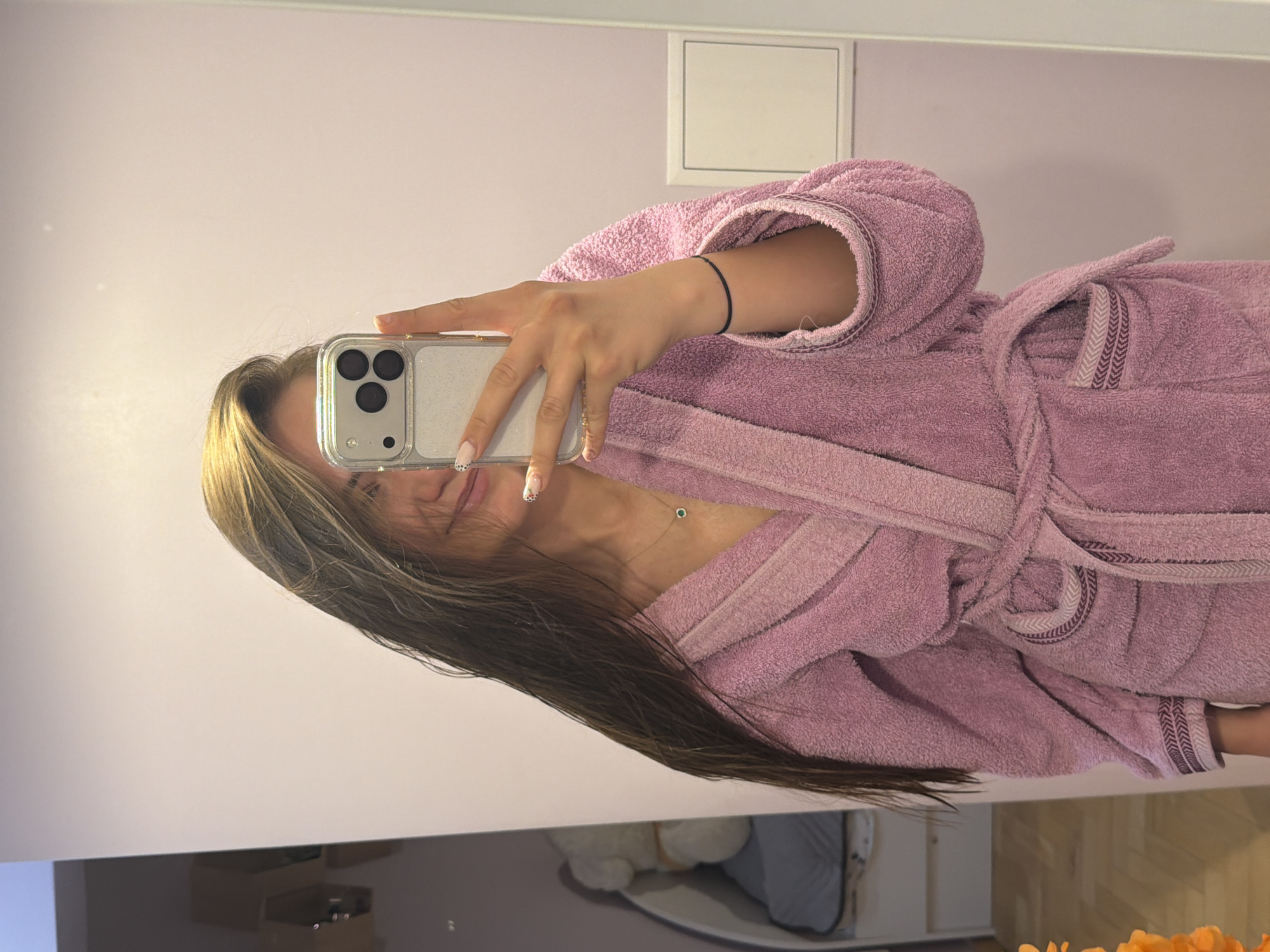 A female individual takes a mirror selfie indoors in Buhovo, Bulgaria. The image, rotated 90 degrees counter-clockwise, captures the person from the chest up. She has long, straight brown hair with lighter highlights.
The subject is wearing a soft, light purple (or dusty rose) terrycloth bathrobe, tied at the waist. The bathrobe features a distinctive striped pattern in varying shades of purple along the cuffs and collar/lapels.
She is holding a smartphone, identifiable as an iPhone due to its triple camera system, encased in a clear, glittery case with a metallic silver frame around the camera module. Her manicured nails are visible, with some featuring white polish and small black polka dots. Accessories include a thin necklace with a small, circular green gemstone pendant and a simple black string bracelet on her left wrist.
The background, reflected in the mirror, shows light purple walls. A white rectangular vent or cover is mounted on the upper wall. The lower reflection reveals blurry indoor furnishings, including what appears to be a white plush object and light-colored wooden flooring. The scene suggests a casual moment, likely in a bathroom or bedroom. No text is visible in the image.