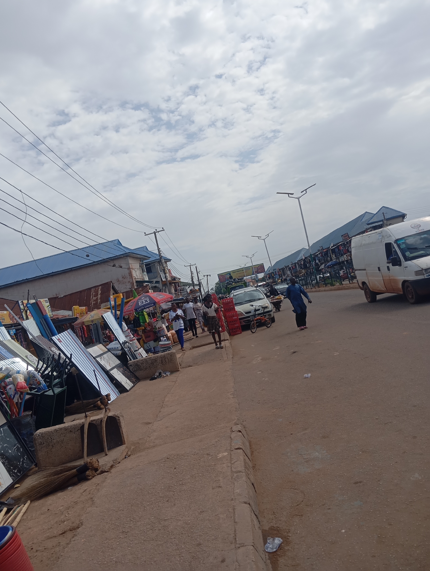 The image depicts a bustling street scene in Bukuru, Nigeria. A street runs through the center of the frame, with a sidewalk on the left. A variety of goods, including metal sheets, tiles, and other hardware items are displayed along the sidewalk, indicating a market or commercial area. People are walking along the sidewalk and street, engaged in various activities. Vehicles, including a white van and a car, are present on the road. The sky is overcast with clouds. Overhead power lines are visible. Street lamps are also seen. The scene suggests a typical day in a busy commercial area in Bukuru.