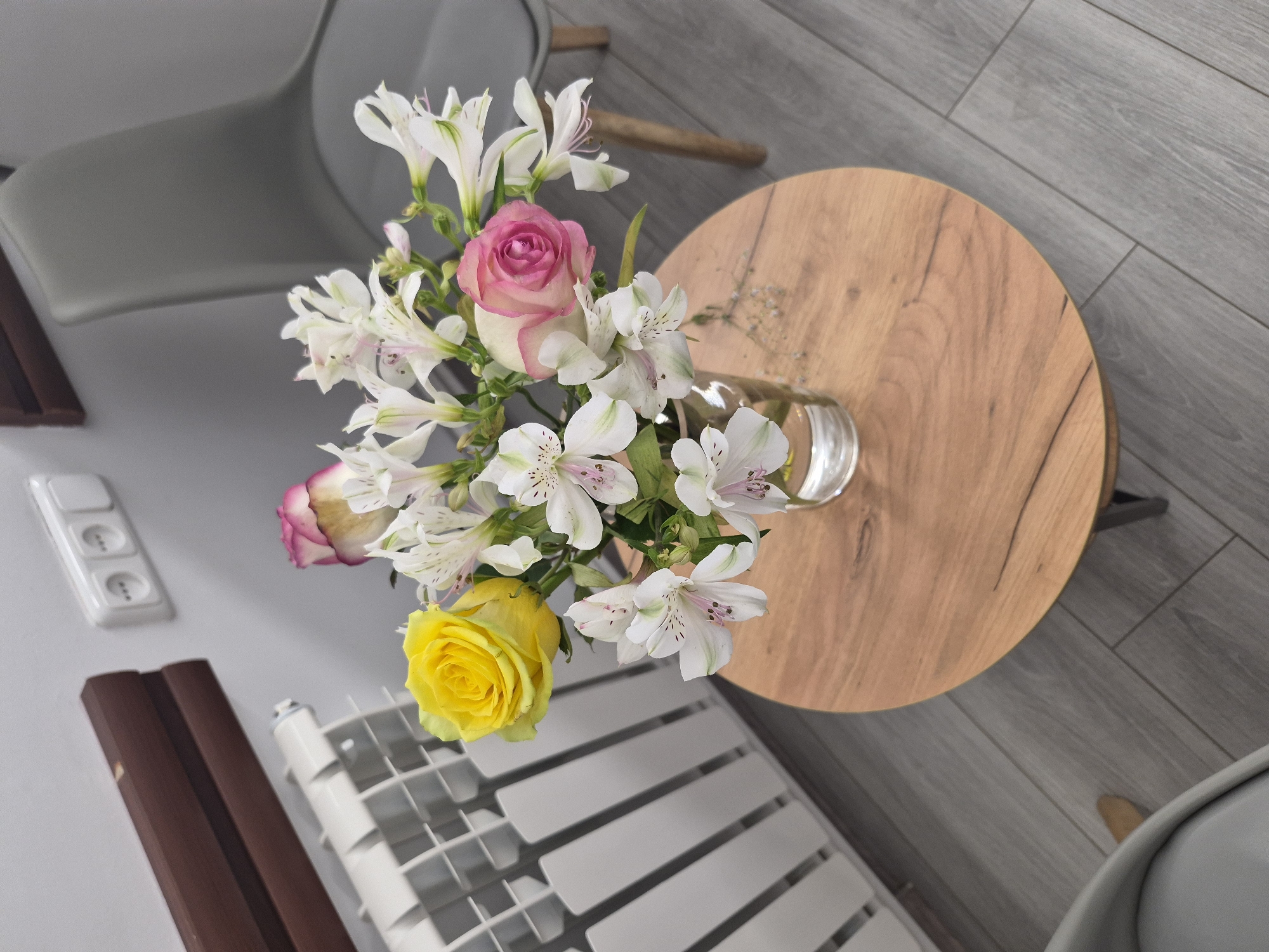 A bouquet of roses and alstroemeria sits in a glass vase on a round wooden table. The bouquet contains two pink roses, one yellow rose, and several white alstroemeria with pink markings. The table is positioned on a gray wood-plank floor. In the background, a white radiator is visible on a gray wall, with a dark brown wooden shelf above it. To the right of the radiator, a white electrical outlet with three sockets is mounted on the wall. A grey upholstered chair is partially visible in the upper right corner of the image, and another grey chair is visible in the lower left corner. The scene appears to be an interior setting, possibly a waiting room or a living space, located in Yerevan, Armenia.