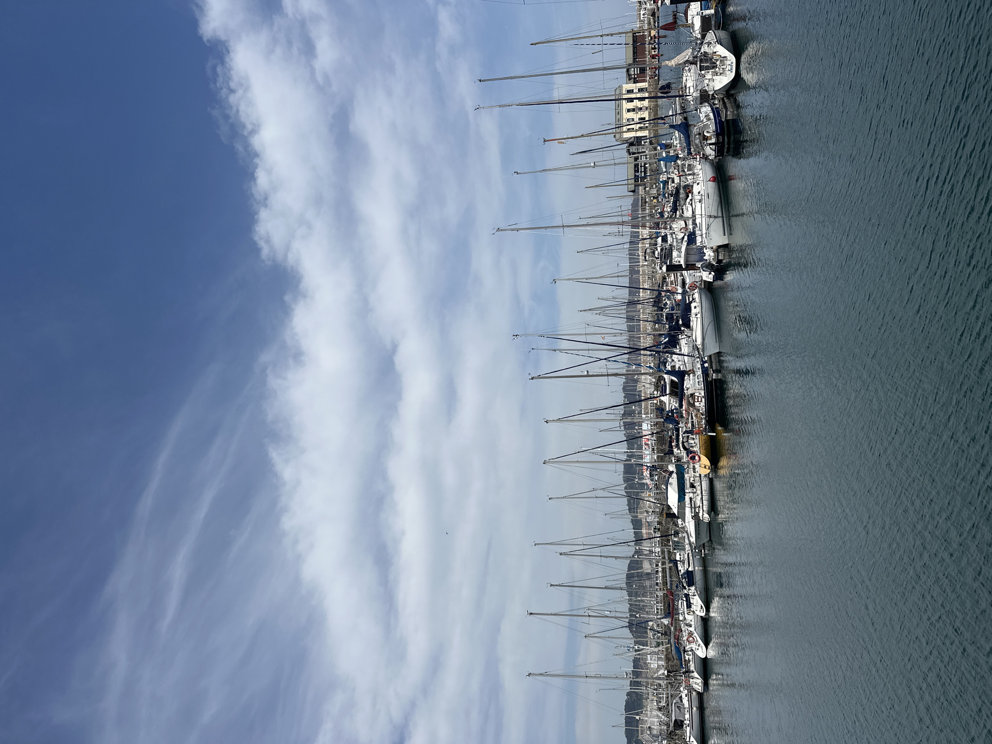 The image depicts a marina with numerous sailboats docked alongside a quay. The masts of the boats create a dense, linear pattern against the backdrop of a bright, blue sky with streaky clouds. The water surface is slightly rippled. Buildings and urban development can be seen along the quay, suggesting a city location. Given the mention of Gijón/Xixón, Spain, this likely depicts the Marina of Gijón. No people are visible in the image.