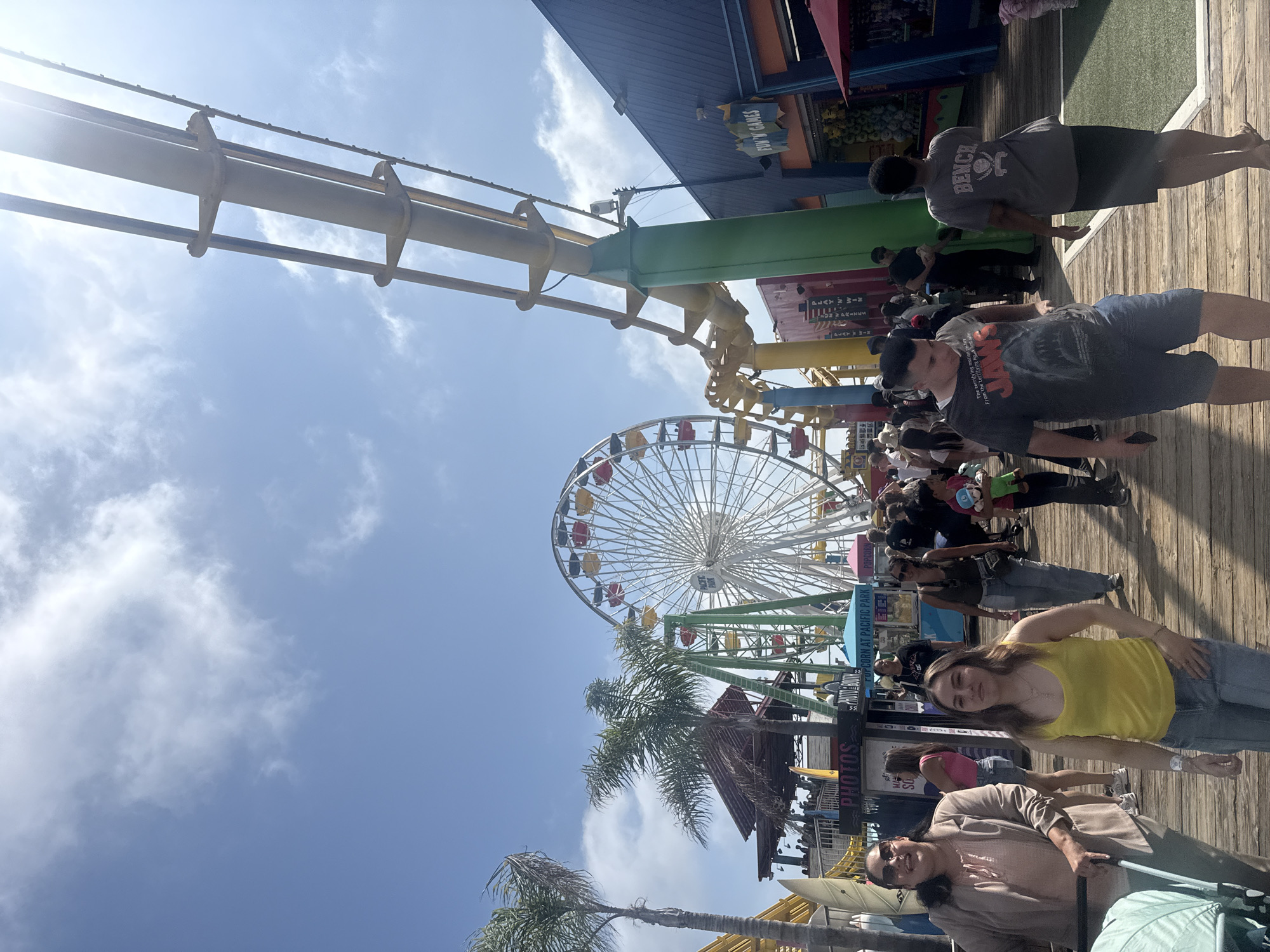 The image depicts a sunny day scene at the Santa Monica Pier in Santa Monica, California, USA. A Ferris wheel, Pacific Park's iconic landmark, dominates the background. Numerous people are walking along the wooden pier. There are palm trees and arcade games. The sky is blue with scattered clouds.