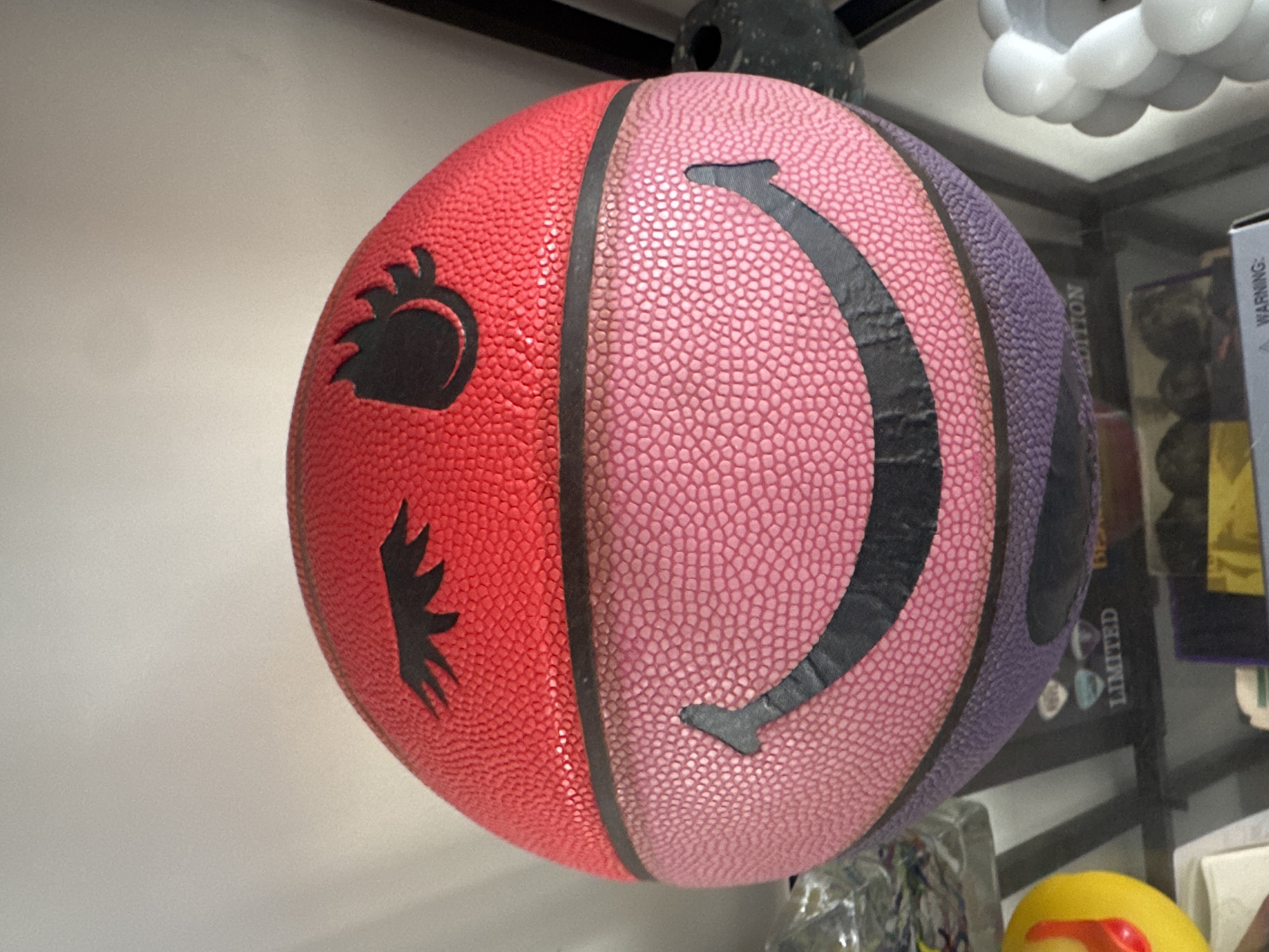 A close-up shot shows a multi-colored basketball, predominantly red, pink, and purple. The basketball features black designs: an abstract, flame-like emblem on the red portion, and a curved line resembling a smile across the pink section. A black stripe separates each color section of the ball. The ball is positioned in a retail setting, possibly a store or display case, as indicated by the visible shelf structure and surrounding items, including a stack of items on the right and a light fixture at the top of the frame. The precise location within London, United Kingdom, cannot be determined from the image.