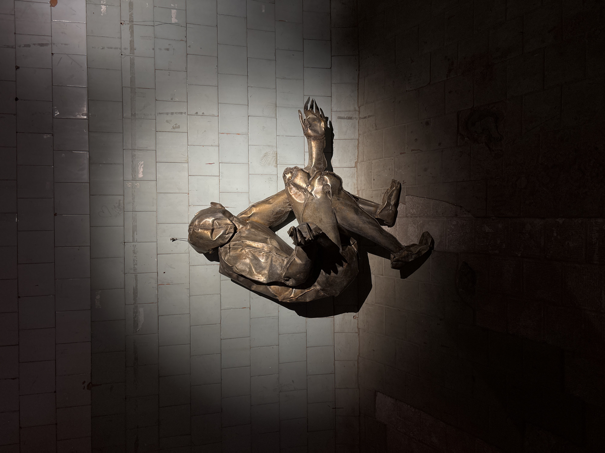 The image depicts a metal sculpture of a human figure, seemingly lying on its side within a dimly lit room. The room is characterized by tiled walls, with the tiling pattern visible both on the floor and walls, suggesting the sculpture is placed within a corner. The sculpture's surface appears dented and folded, giving it a raw or unfinished aesthetic. Light is focused on the statue and the corner to the left of the statue; the right side of the statue is significantly darker.
Although I lack explicit details on the city of Saint Petersburg, Russia, or other elements that would support more context around the specific location, the presence of the sculpture gives a hint as to where the piece might be located.
