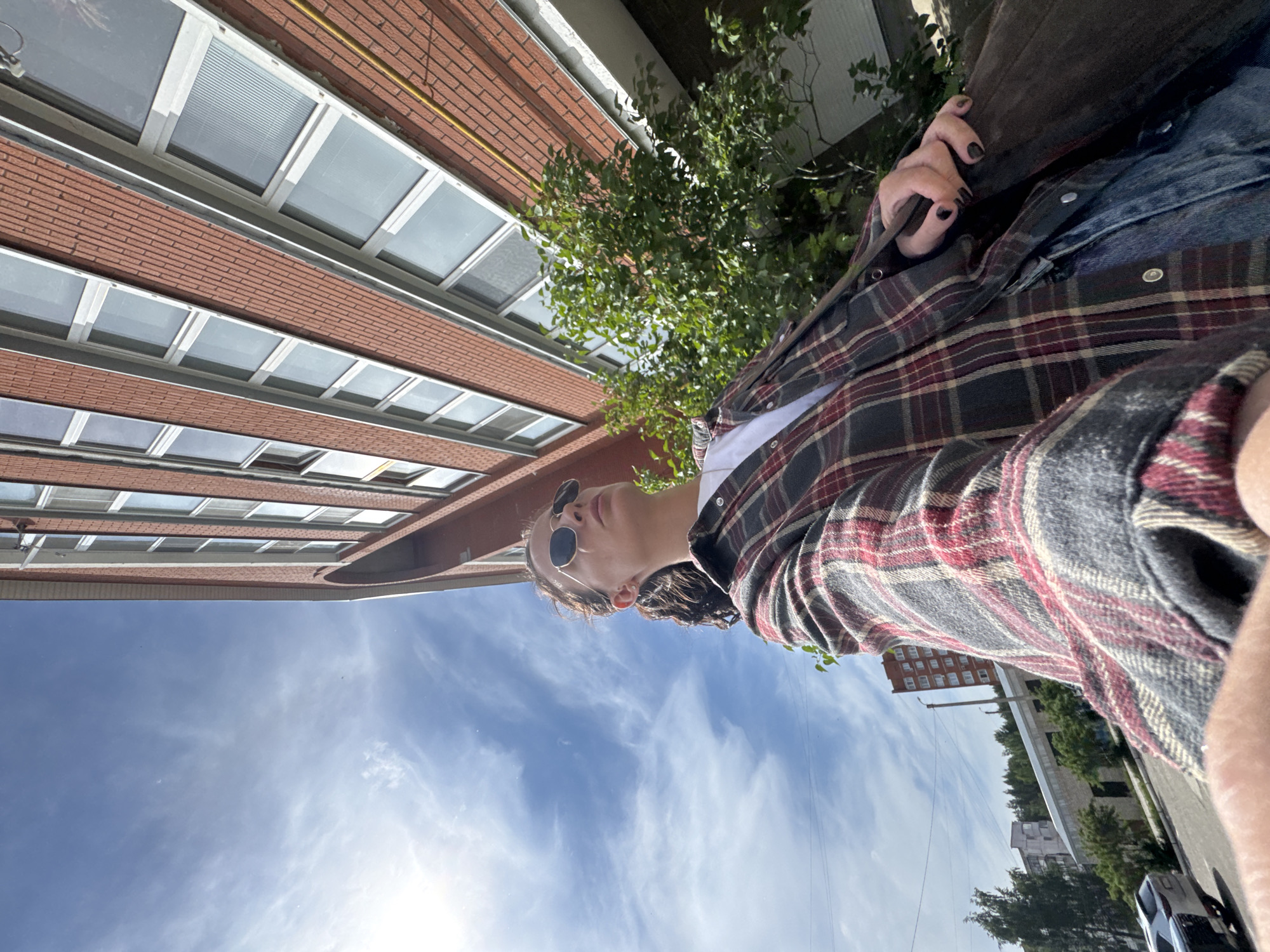 The image, rotated 90 degrees clockwise, captures a person from a low-angle perspective in an urban outdoor setting in Chaikovsky, Russia, during daytime. The individual, likely young, is viewed from the right side, looking slightly upwards. They are wearing dark round sunglasses, a plaid button-up shirt in shades of red, grey, and black over a white top, and a denim layer underneath. Their hair appears to be styled in dreadlocks or braids. One hand, adorned with dark nail polish, is visible, seemingly holding a strap or object.
The background on the left is dominated by a multi-story red brick building with numerous rectangular windows, some featuring blinds, and a yellow pipe running along its upper facade. A green tree with dense foliage is positioned behind the person. To the right, further back, an urban street is visible with other buildings, including one with many small windows and another with a curved structure, along with utility poles and overhead wires. A silver car can be seen on the street.
The sky is bright blue with scattered white, wispy clouds, indicating clear and sunny weather. The lighting suggests it is mid-day or afternoon. There is no visible text in the image.