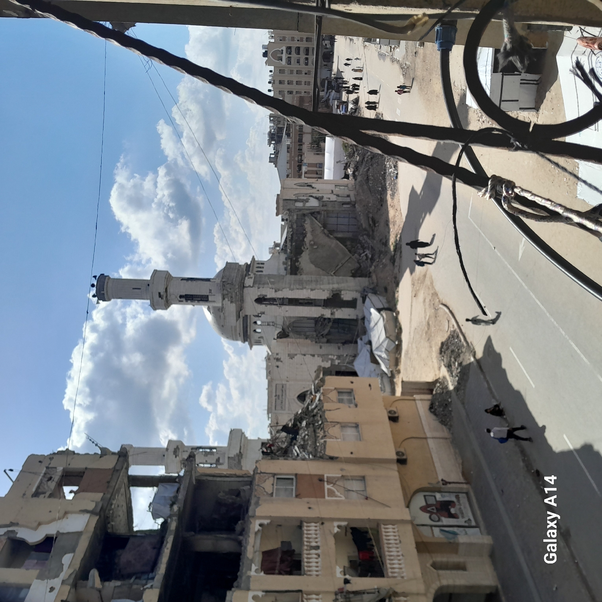 The image shows a city street with buildings that are severely damaged or destroyed. A mosque with a tall minaret and dome stands in the center of the frame, showing signs of damage. People are seen walking along the street. Cables and wires are visible in the foreground, possibly obscuring the view. The sky above has clouds and blue patches. "Galaxy A14" is printed on the bottom right.