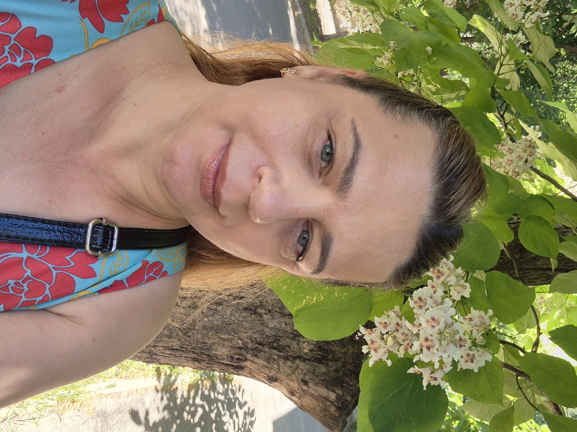 A medium shot portrait shows a woman in her 40s with light brown hair pulled back, wearing a light blue and red floral dress. She is standing outdoors, with a tree trunk and branches with white flowers and green leaves in the background. Her left arm is visible, and she is wearing a gold earring. A black purse strap is visible on her right shoulder. The lighting suggests it is daytime, and the background indicates an urban or suburban setting with a road and grass visible.
