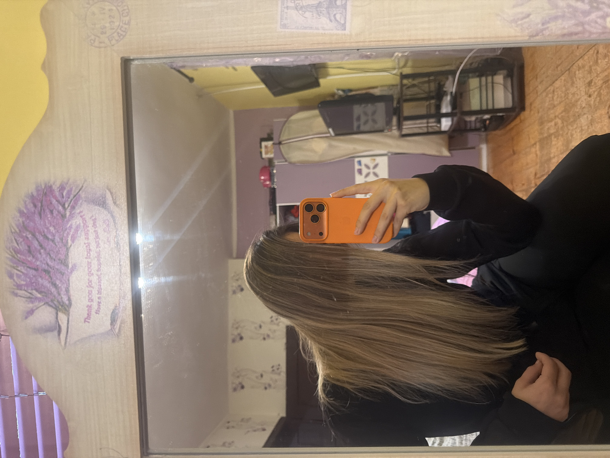 The image shows a woman taking a selfie in a mirror. The mirror has a wooden frame with a floral design on the left side. The woman is holding an orange phone in front of her face, obscuring her features. She has long, light brown hair and is wearing a black long-sleeved shirt.
The background reflected in the mirror shows a room with light purple walls and white patterned wallpaper. There is a TV on a shelf, various household items, and a curtain visible.
The location is Unknown, Unknown.