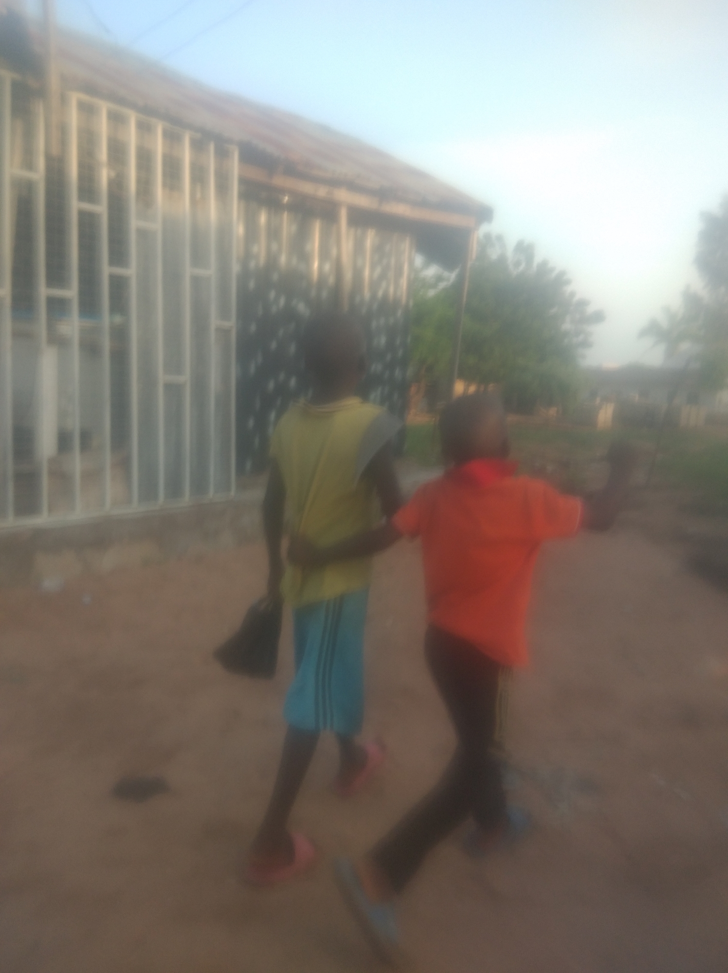 Two young boys are walking away from the camera on a dirt path in front of a building in Jalingo, Nigeria. The boy on the left is wearing a yellow sleeveless shirt and blue shorts, and he is carrying a black bag in his right hand. The boy on the right is wearing an orange t-shirt and dark pants. The building behind them appears to have a corrugated metal roof and white bars over its windows. Trees and other structures are visible in the background. The sky is clear and blue.